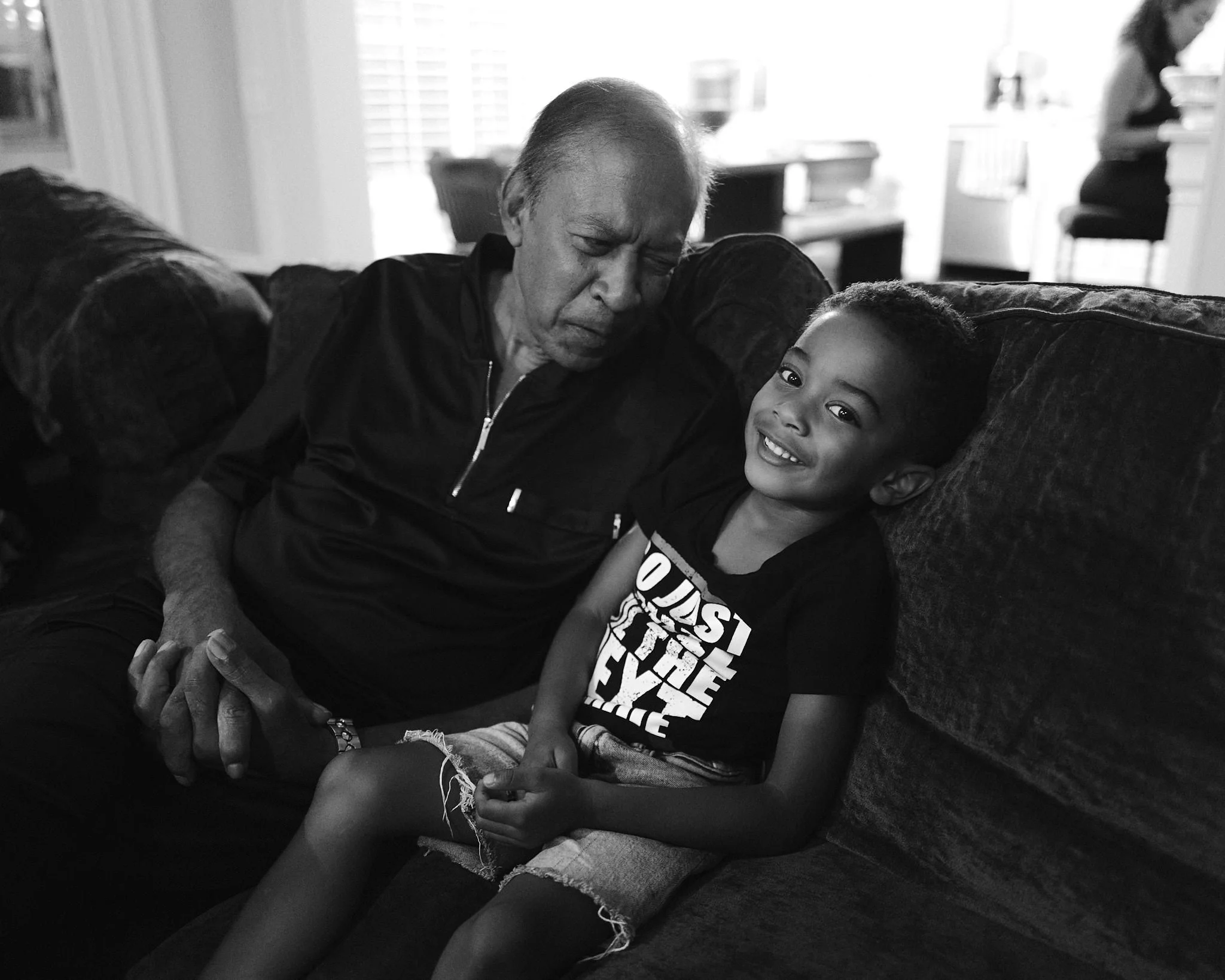 An elderly man and young boy sitting on a sofa, sharing a moment of connection. The boy is smiling and looking at the camera, while the man looks down with eyes closed. The background includes a person sitting at a table.