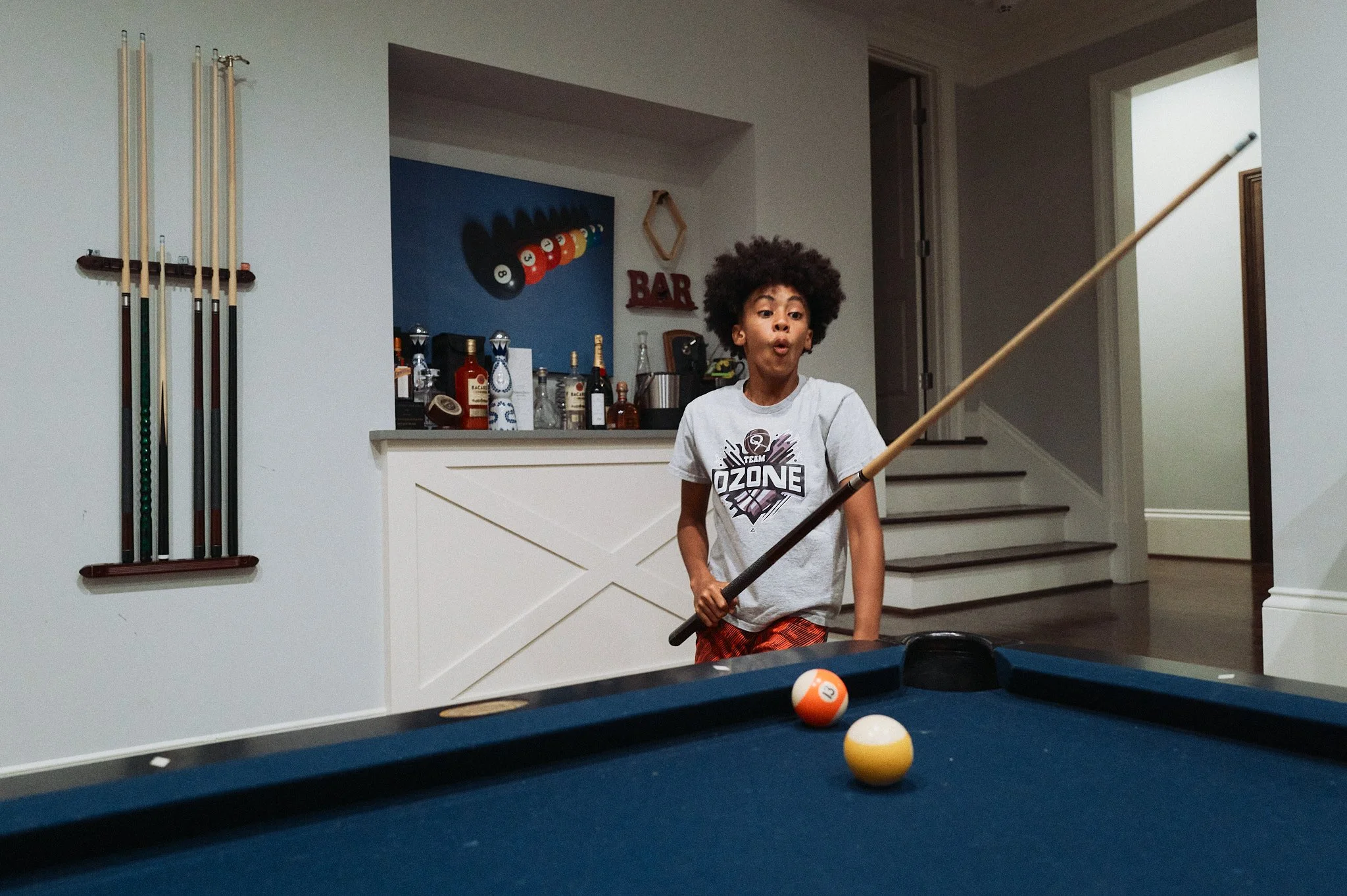 A young boy with an afro hairstyle holding a pool cue, playing pool in a game room, with billiard balls on the table in front of him.