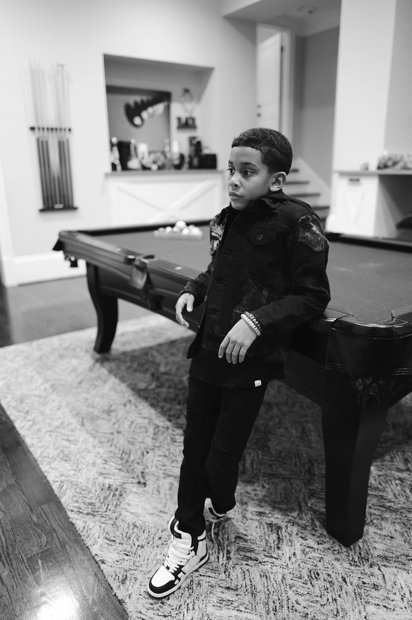 A young boy standing next to a pool table in a living room, wearing a dark jacket and sneakers, with a contemplative expression.