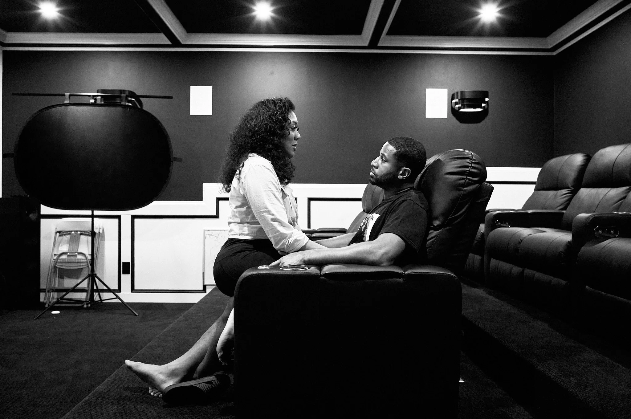Devale and Khadeen Ellis sitting on a leather couch facing each other, holding hands and gazing into each other's eyes, in a dark room with theatre-style seating and professional lighting equipment.
