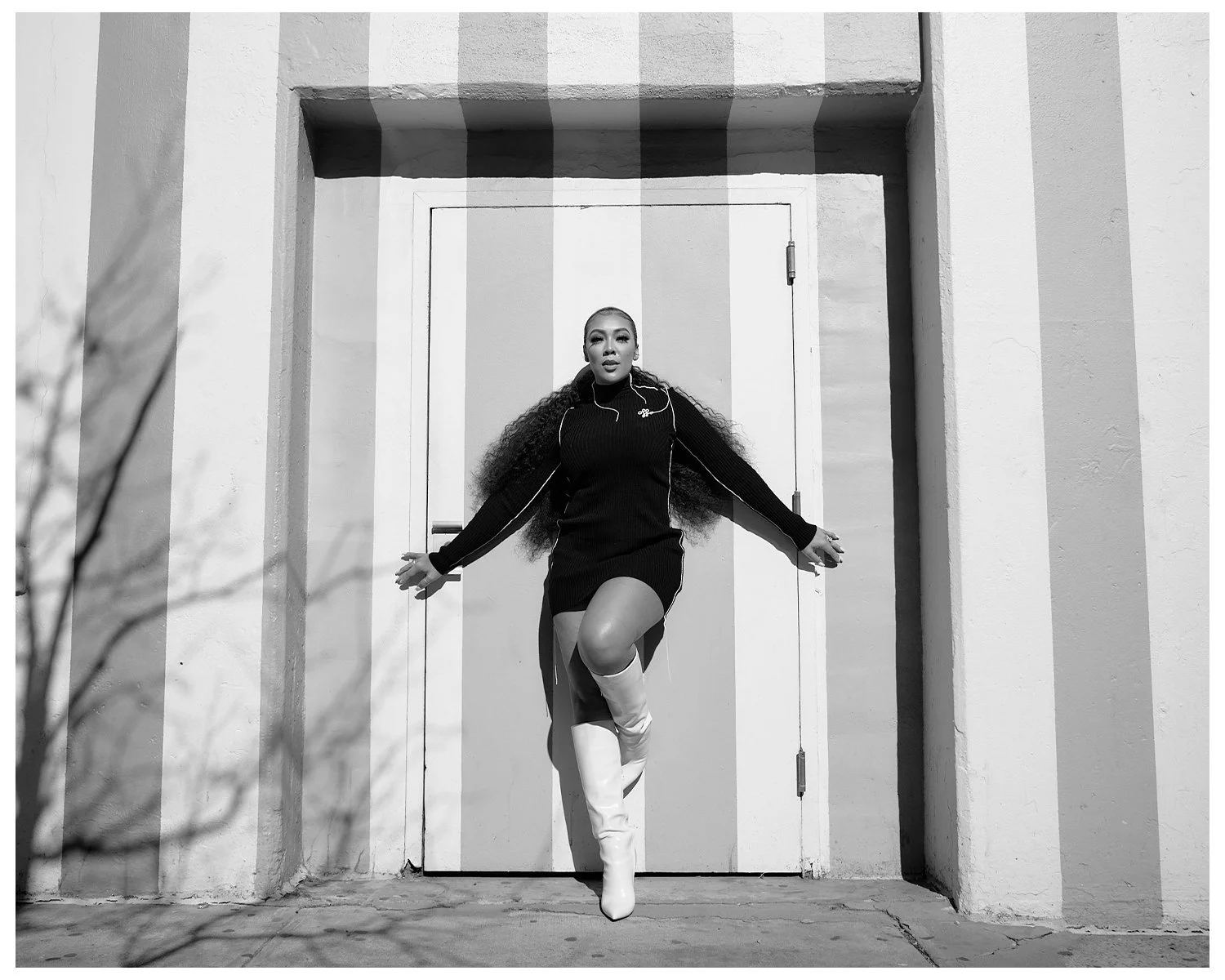 A woman with long curly hair wearing white thigh-high boots and a black outfit with zipper details, striking a pose with arms outstretched in front of a closed door.