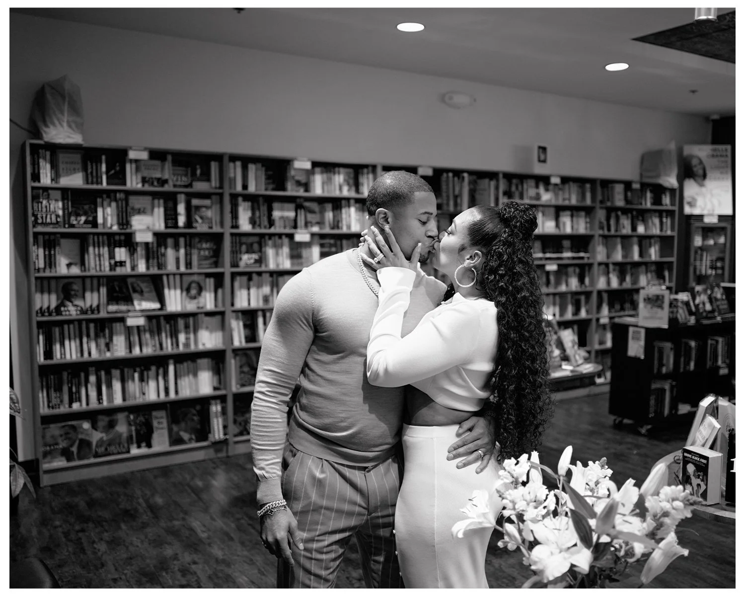 The Ellises kissing in a bookstore, with the woman wearing large hoop earrings and a white outfit, and the man in a long-sleeve shirt and striped pants, surrounded by bookshelves and flowers.