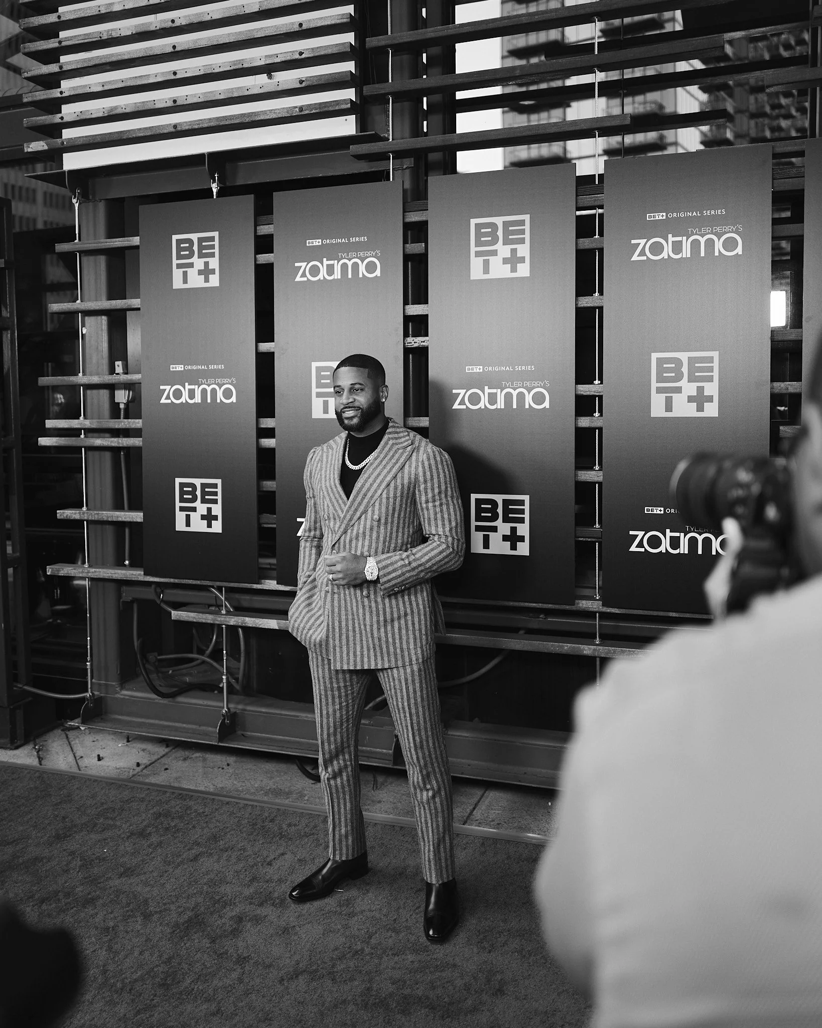 Devale Ellis in a striped suit standing in front of promotional banners for BET+ and Tyler Perry's 'Zatima' at an event, with someone filming him in the foreground.
