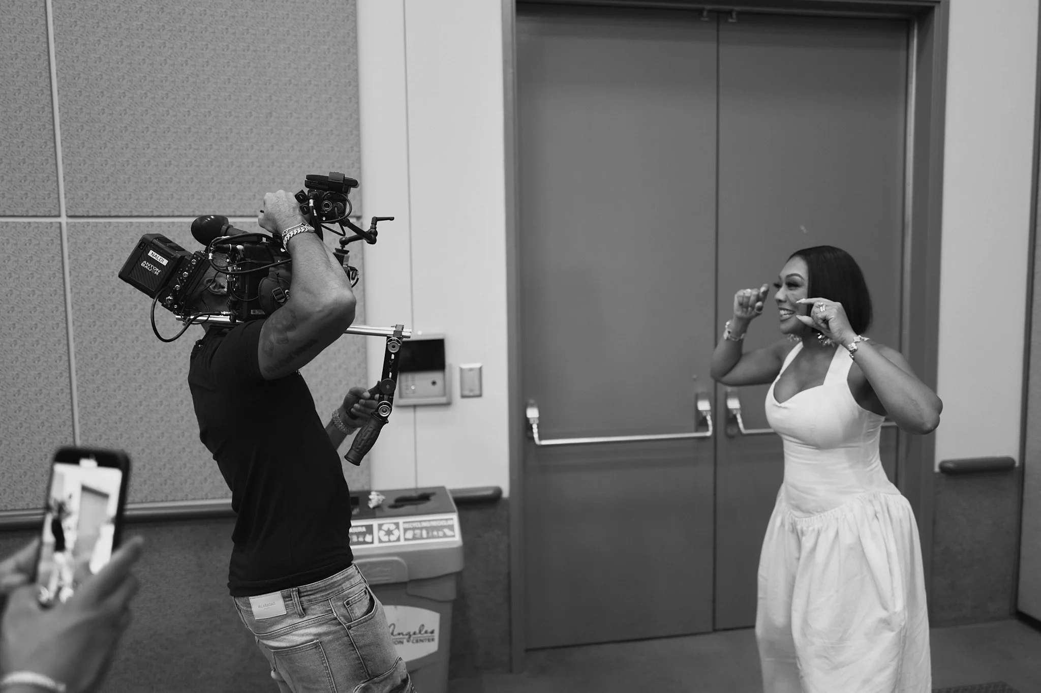 A woman in a white dress smiling and posing for a camera as a man behind the camera records her in a room with a closed door and a trash bin.