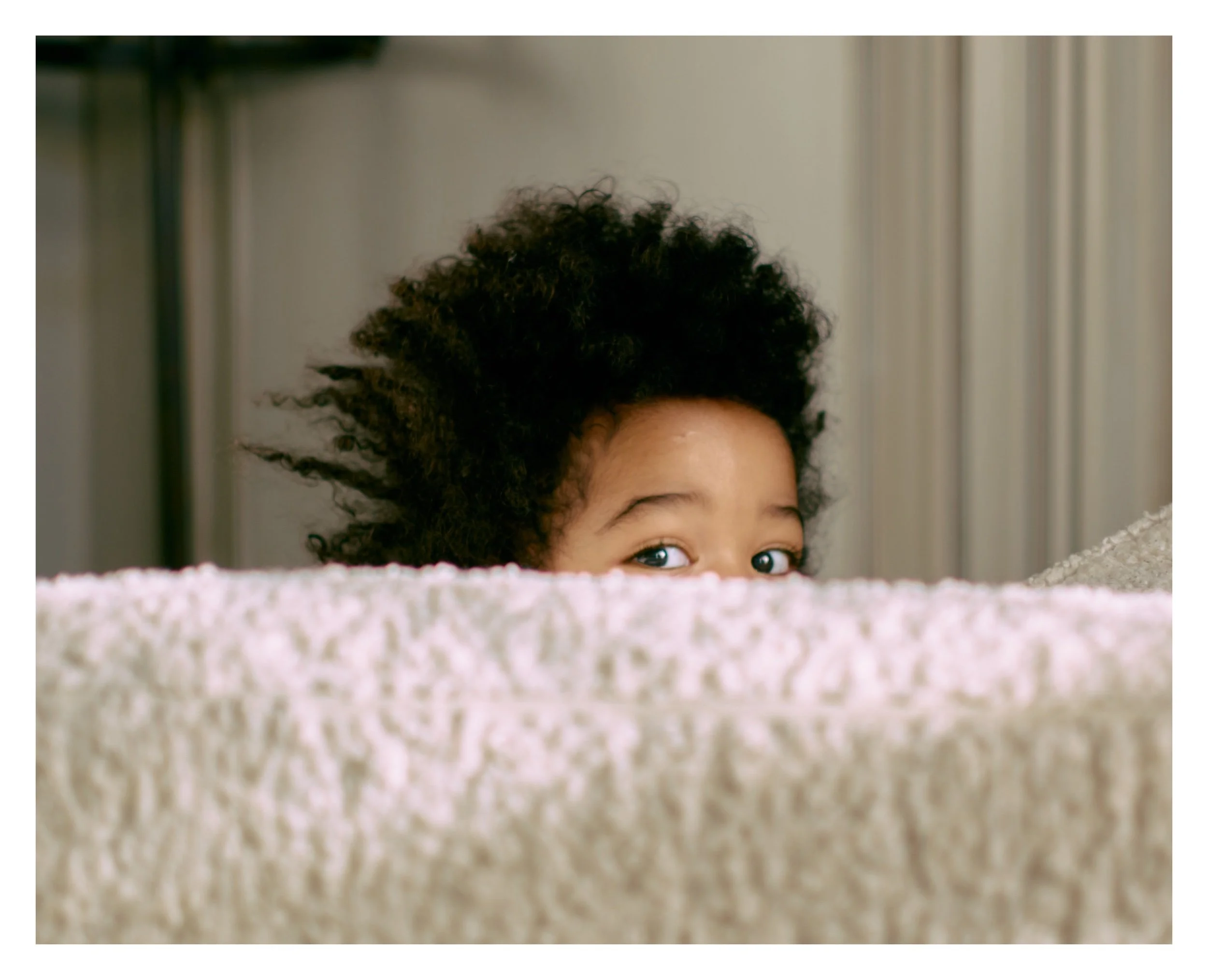 A child with curly black hair peeking over a beige textured surface, with only their eyes and a portion of their forehead visible, in a soft indoor setting.