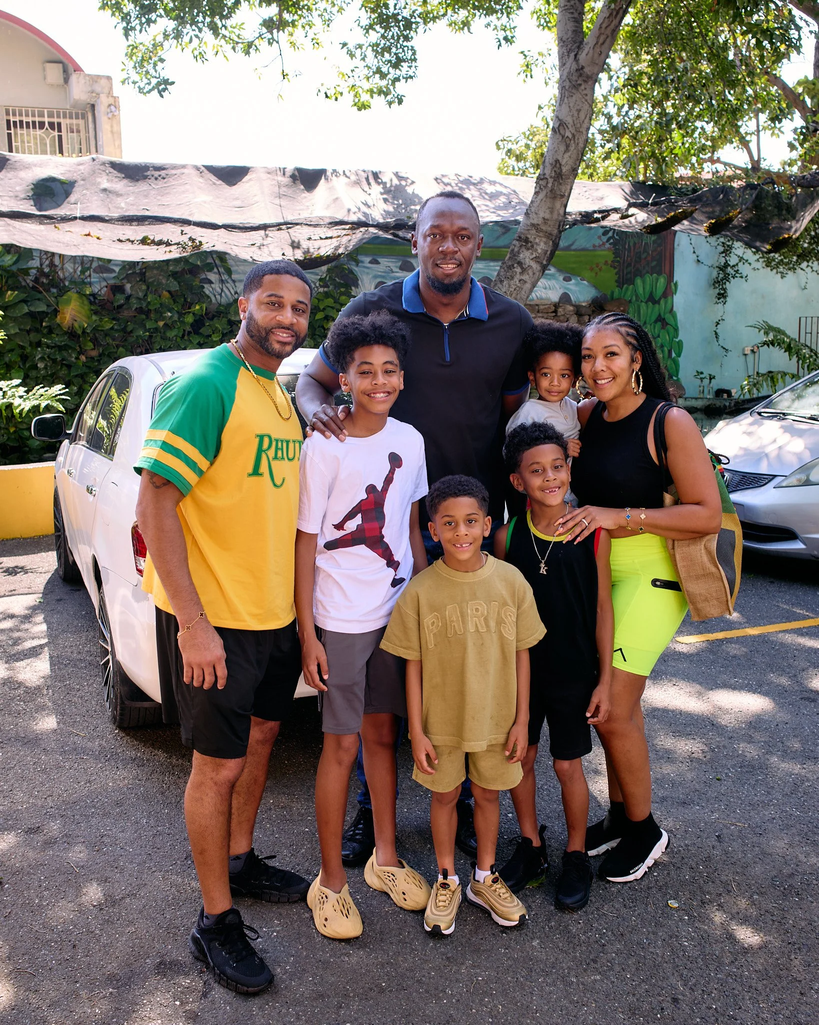 Group of eight people, including Usain Bolt, Devale and Khadeen Ellis standing outdoors in a parking lot with cars and greenery, smiling at the camera.