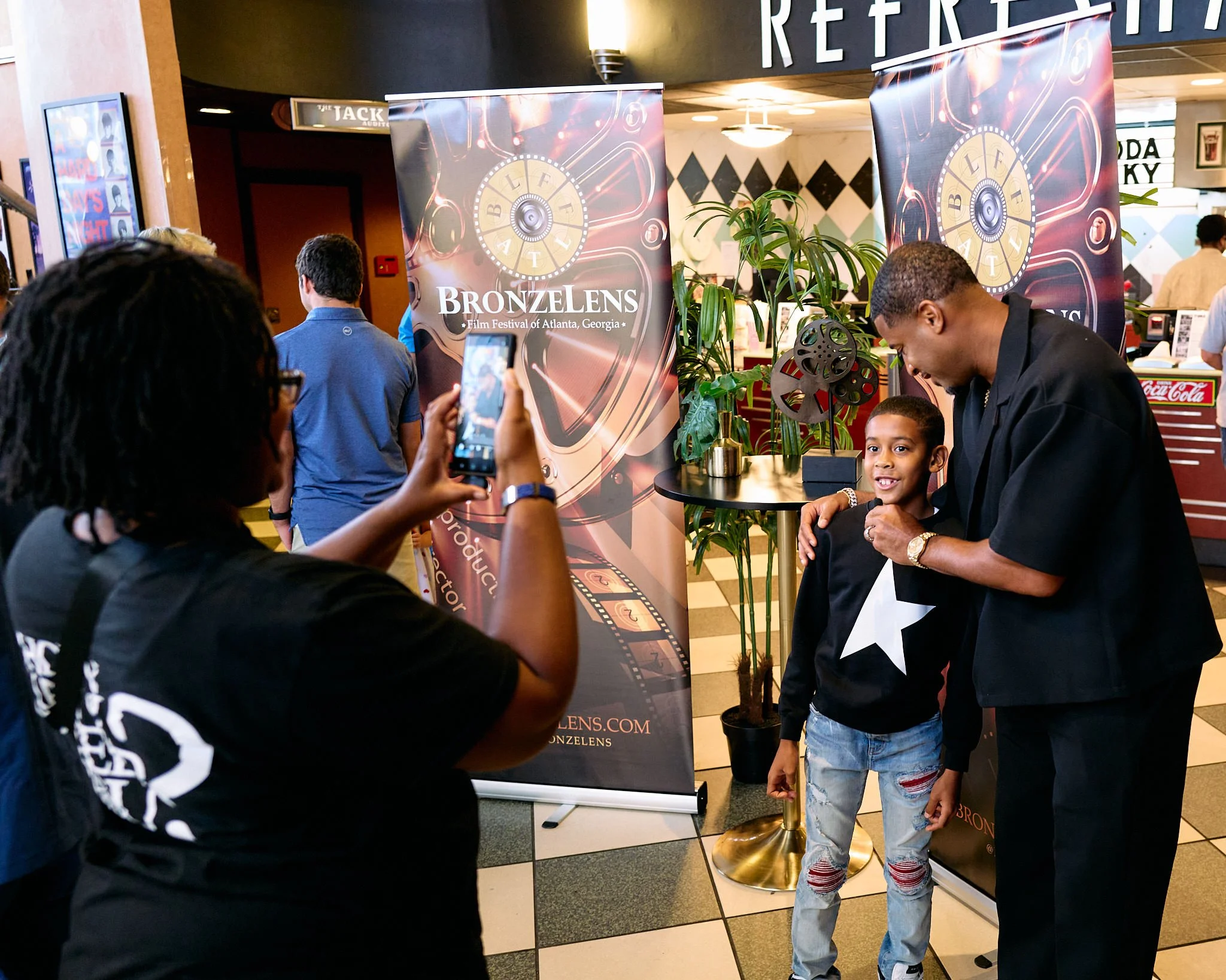 A group of people at an event, with a young boy being photographed by a man. A woman in the foreground is taking a photo of the boy and a man. There are banners with the text 'BRONZELENS' and 'Film Festival of Atlanta, Georgia' in the background.