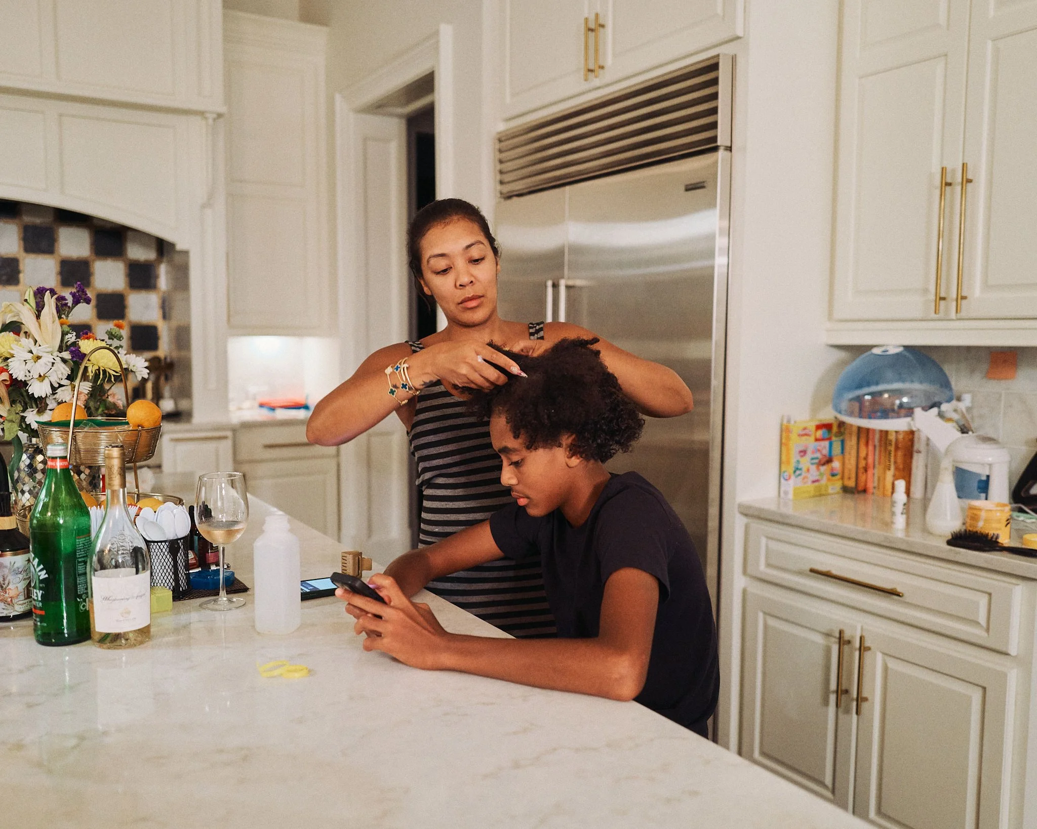 A woman is brushing a young person's hair in a kitchen with white cabinets, a marble counter, and various items on the counter including bottles, a glass of wine, and flowers.