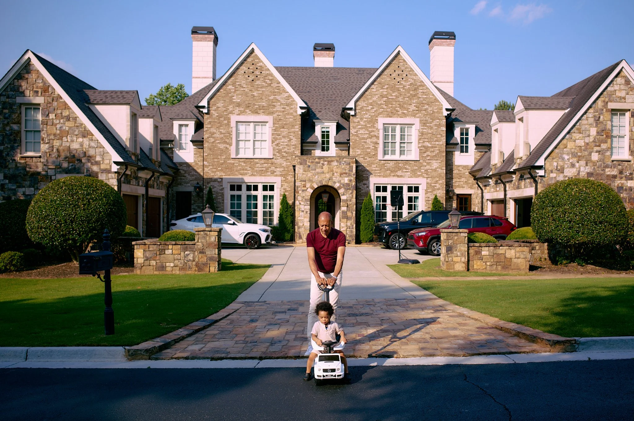 A man and a young girl playing with a toy truck in front of a large brick house with multiple chimneys and parked cars, green bushes, and a driveway.