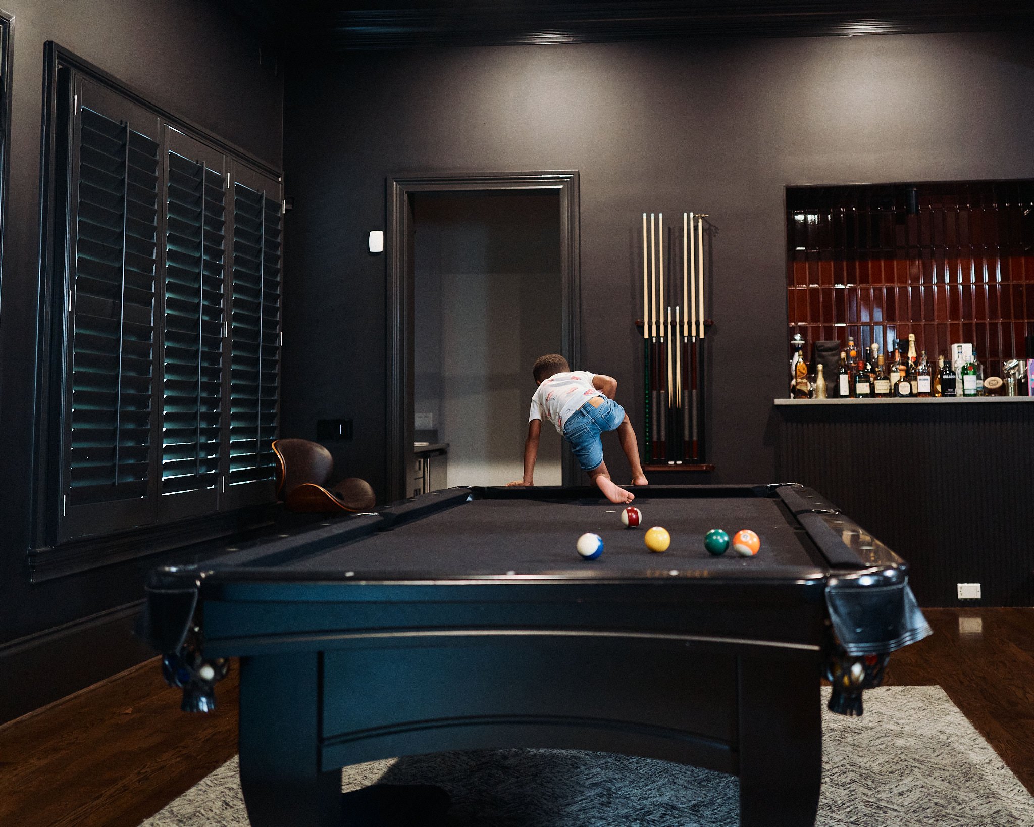 A young boy is playing pool on a black billiards table in a dimly lit game room. There are billiard cues on a rack on the wall and a bar with various bottles of alcohol in the background.
