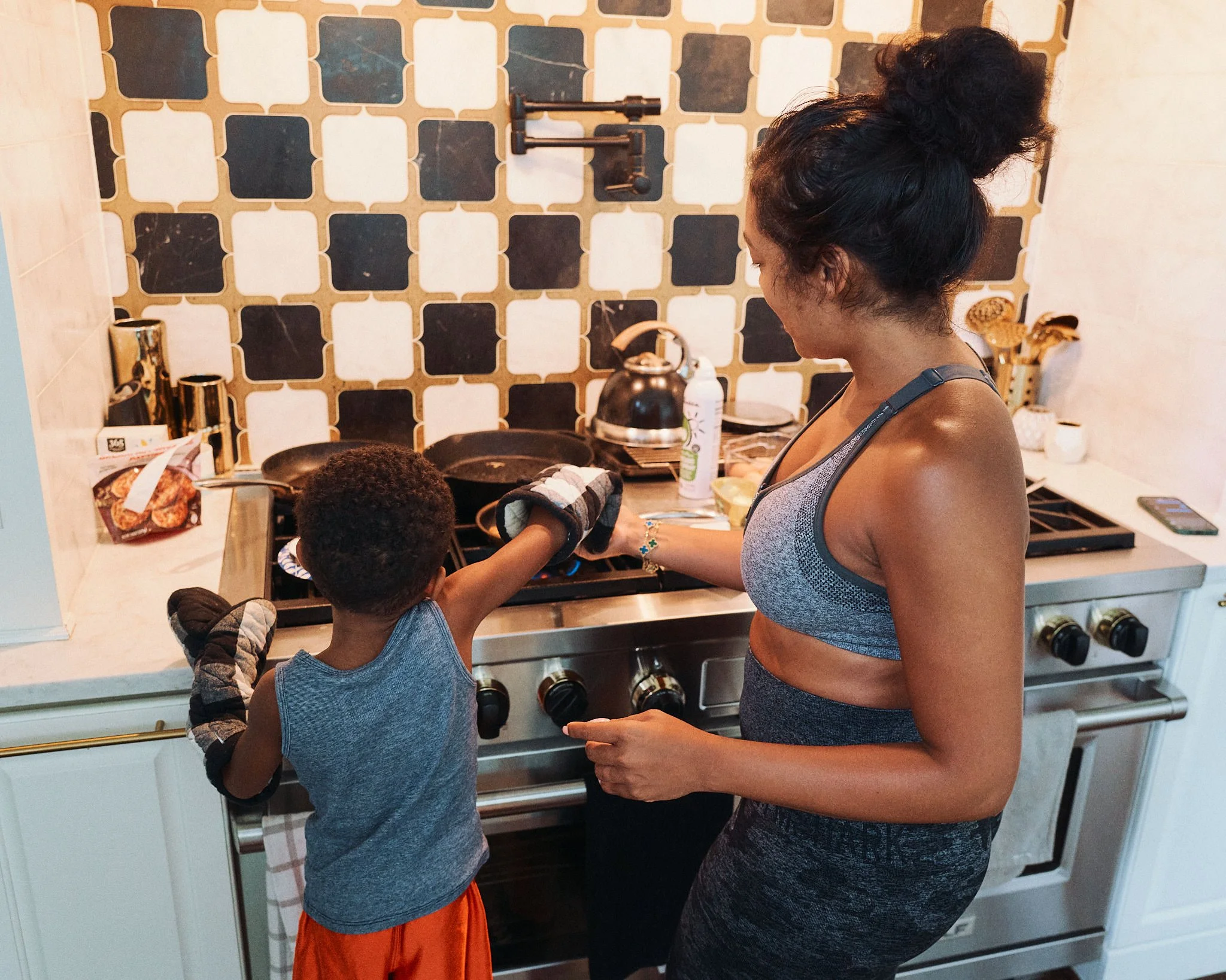 A woman and a young boy cooking together in a kitchen. The boy is reaching for something on the stove while wearing oven mitts. The woman is assisting him, wearing athletic clothing. The kitchen has a patterned tile backsplash and various kitchen ute