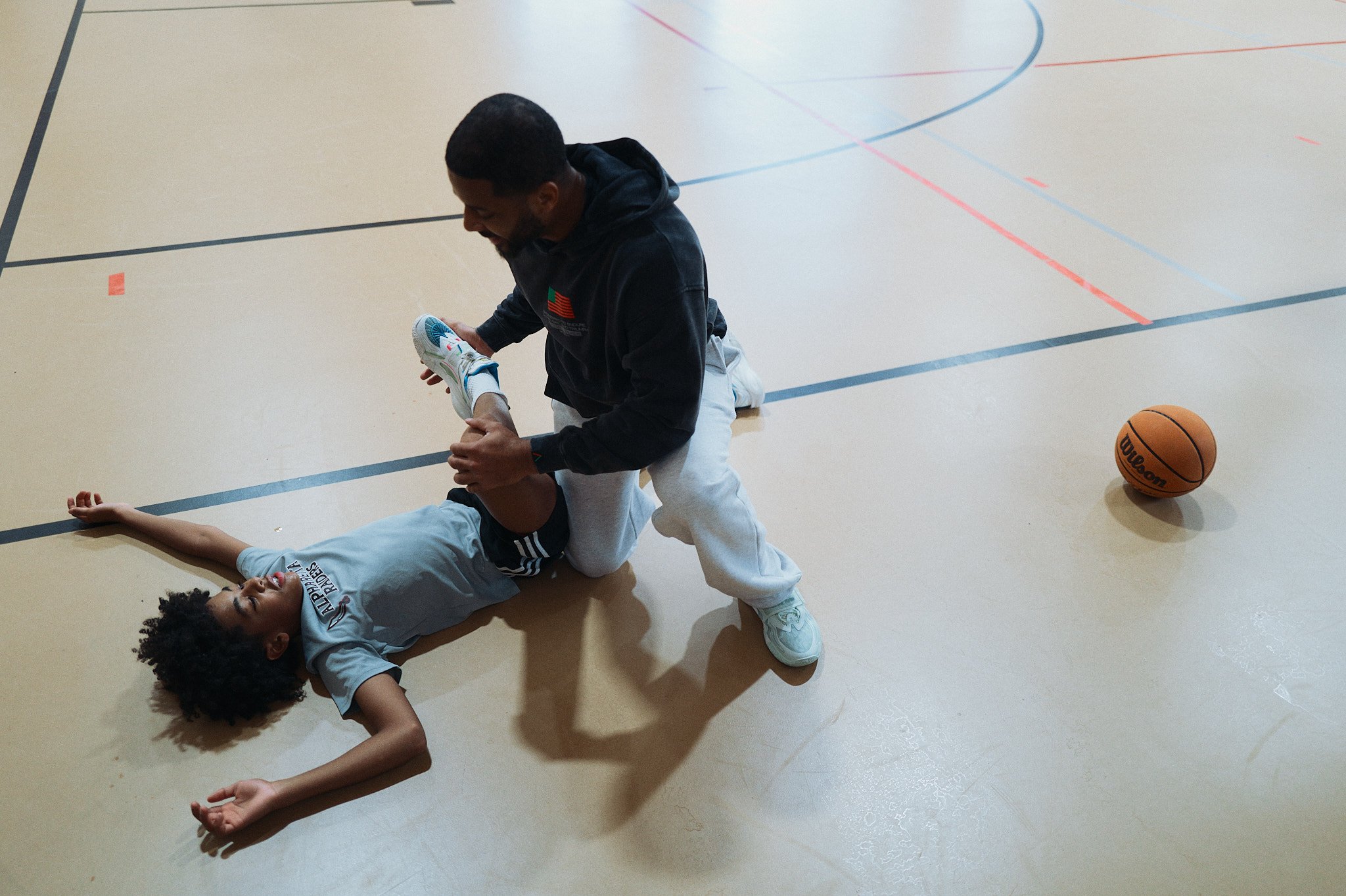 Devale Ellis helping a Jackson Ellis on the basketball court. The girl is lying on her back on the floor, holding her arm, while the man is kneeling beside her, assisting her with her shoe. A basketball is on the floor nearby.