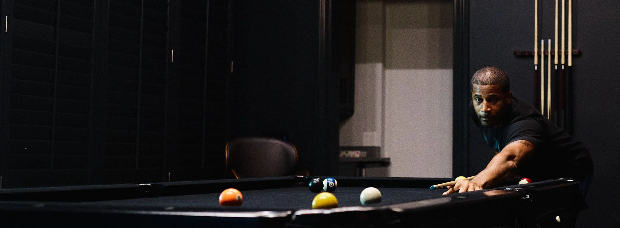 A man with short hair wearing a black shirt playing pool in a dimly lit room, aiming to hit the balls on the black pool table.
