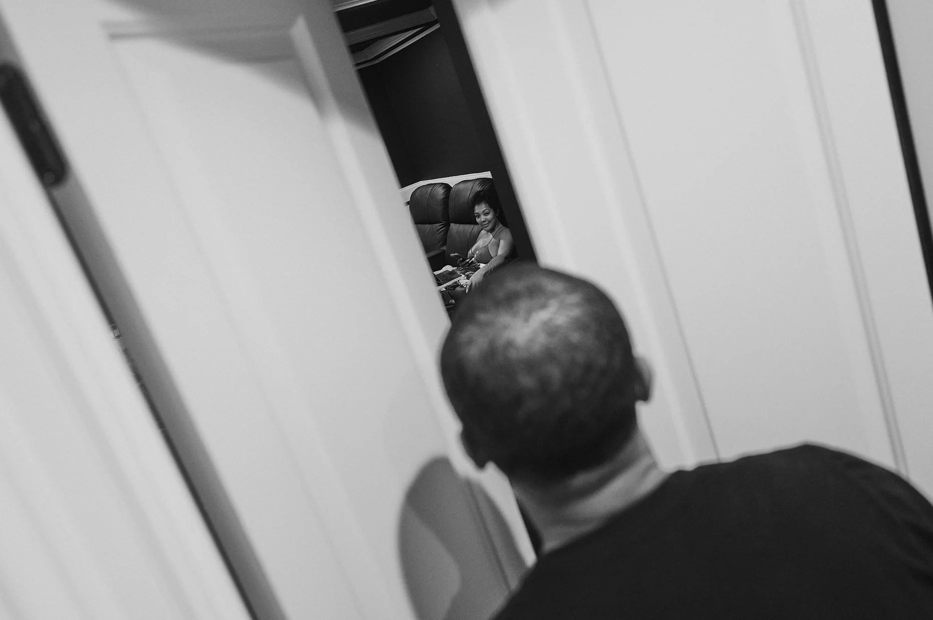 A black and white photo of a Devale looking into a room, seeing a Khadeen seated on a couch and smiling, with her hair styled, in a room with dark walls.