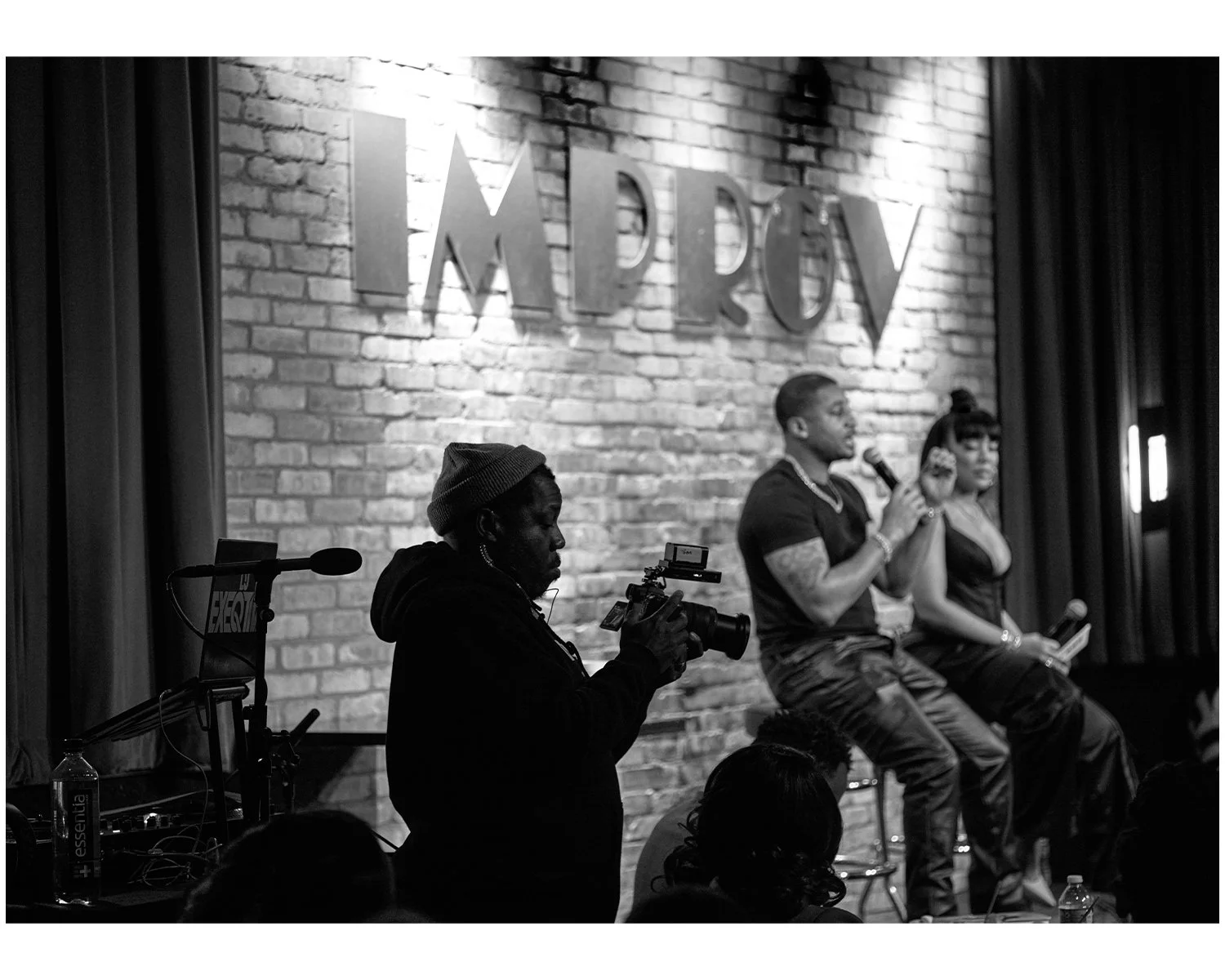A black and white image of a panel discussion at a venue called 'Impact.' Two women and one man are seated on a stage, with the man speaking into a microphone. A cameraman is recording the event, and there are audience members in the foreground.