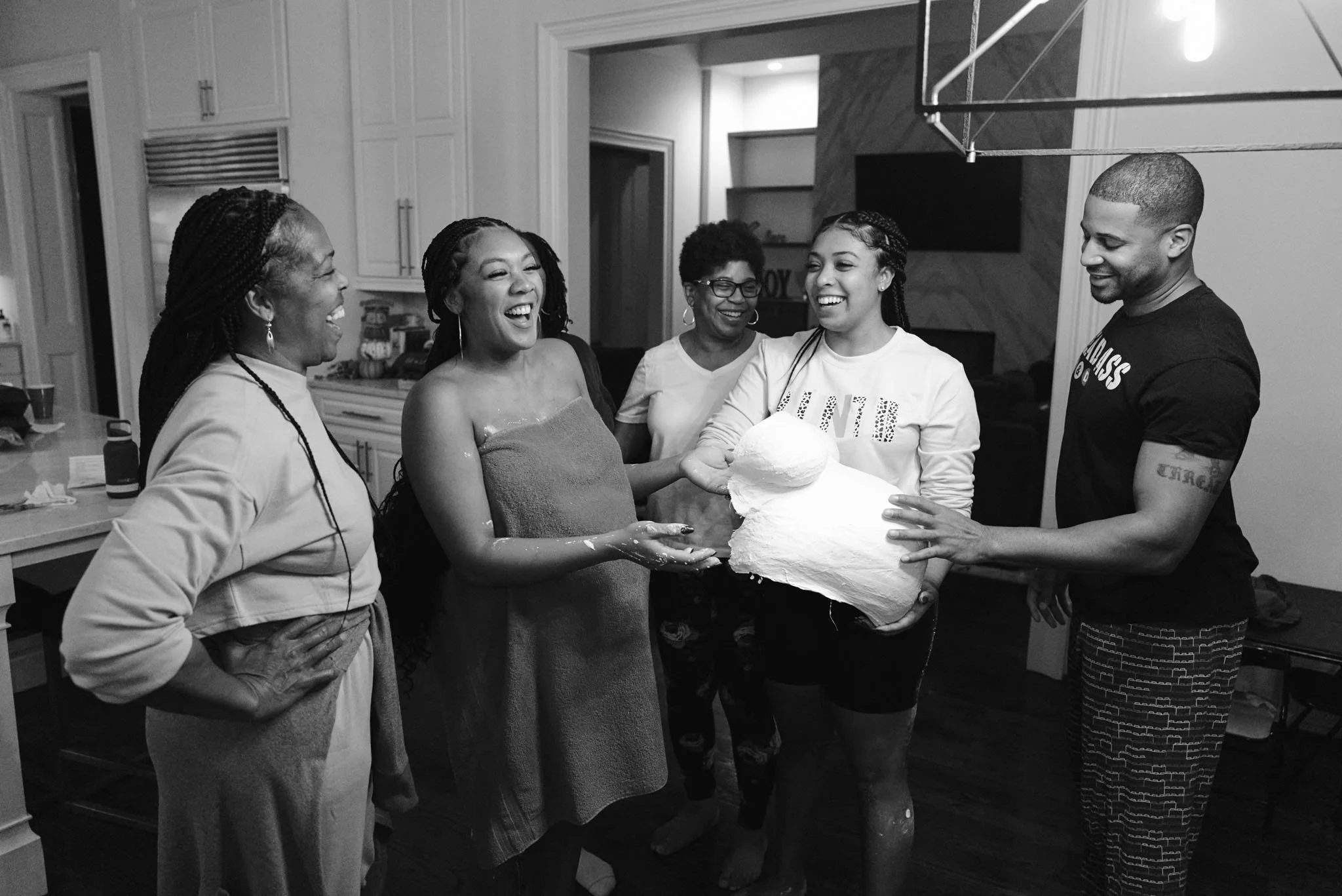 Group of five people smiling and laughing inside a house, participating in a birthday celebration with a cake made of foam or some other material.