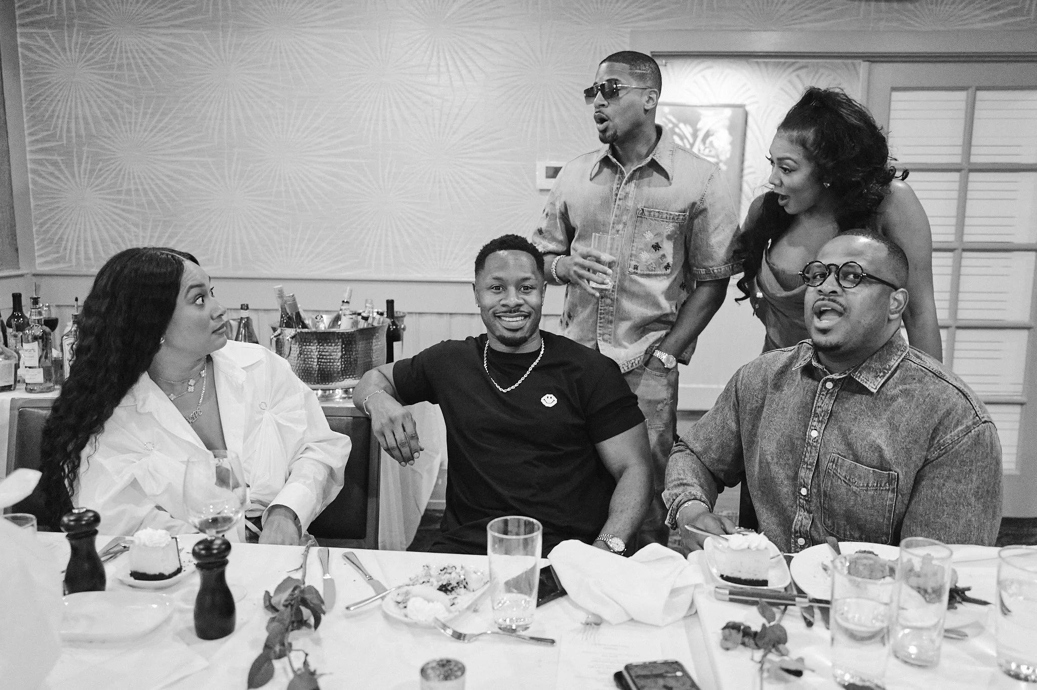 Group of five people sitting and standing around a dining table, engaging in conversation, with plates of food and drinks, at a social gathering or party.