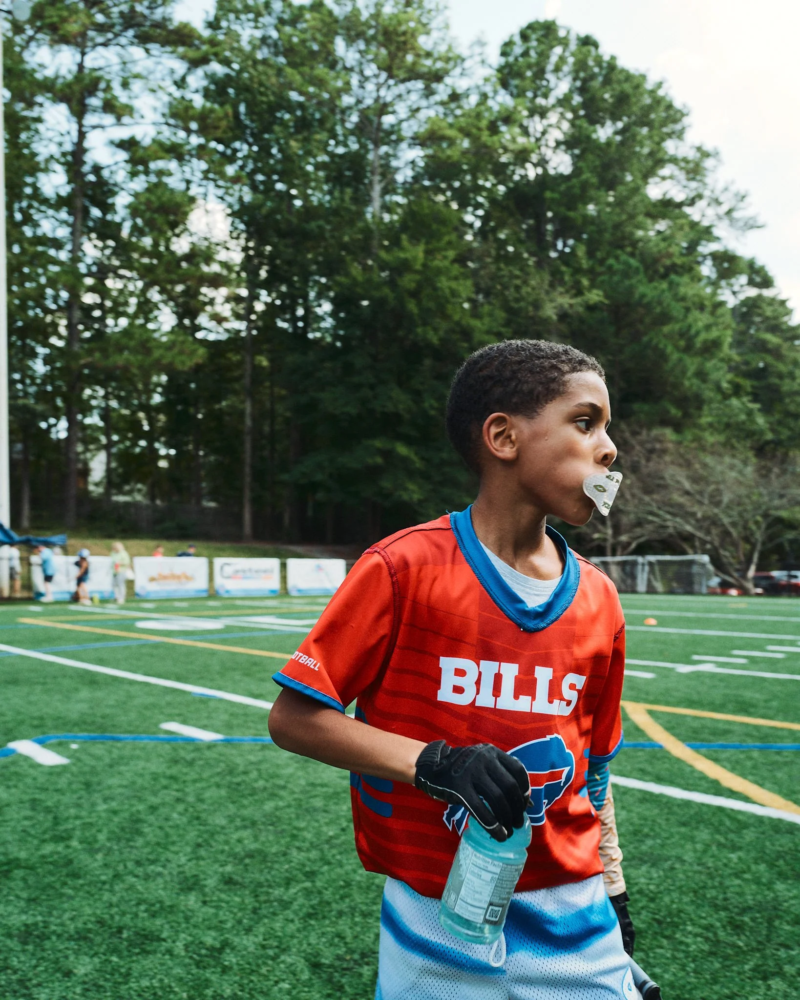 Young boy in a red football jersey with 'BILLS' on it, holding a water bottle, running on a football field with trees in the background.