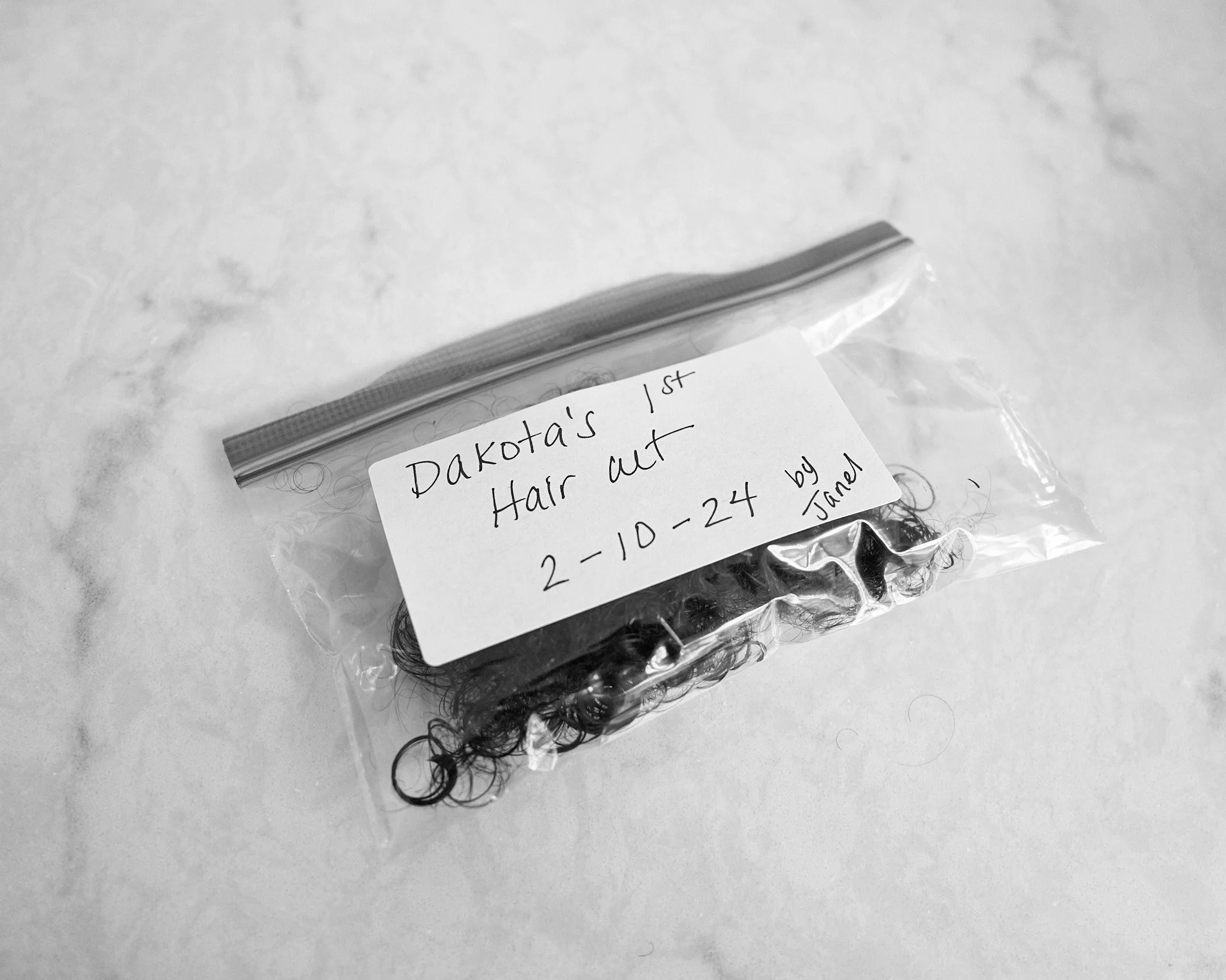 A small plastic bag containing dark curly hair labeled with handwriting that reads 'Dakota's 1st Hair out 2-10-24 by Janet'.