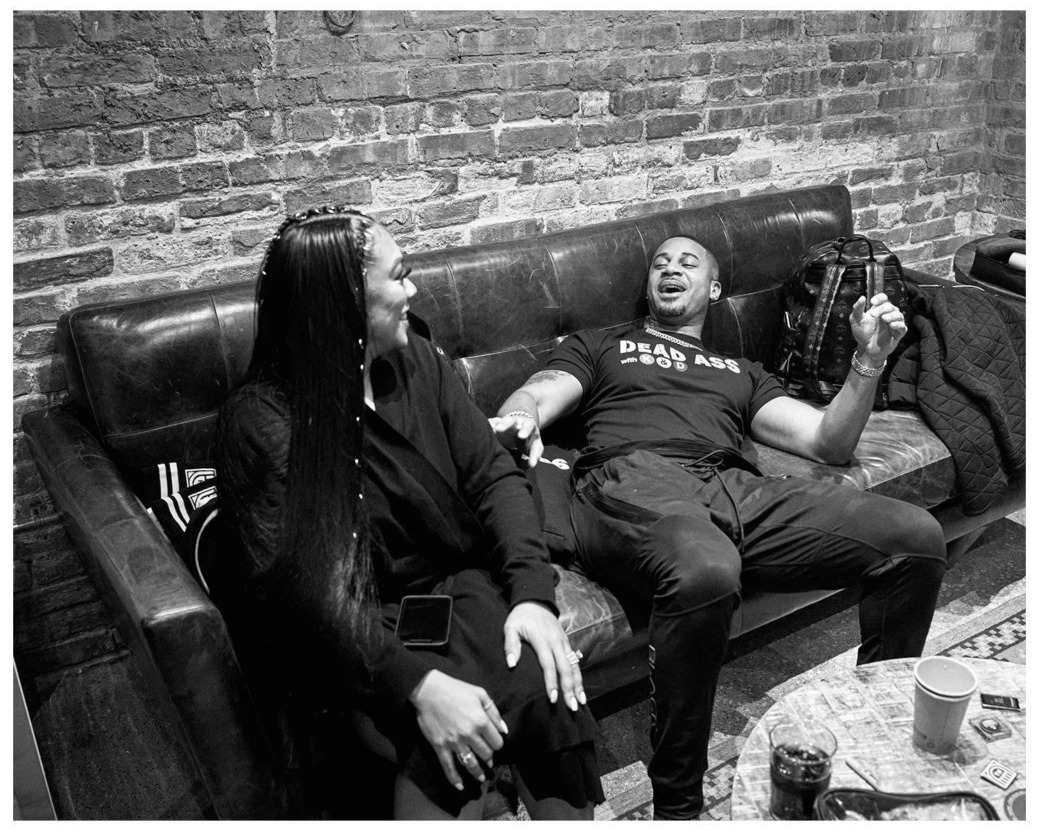 A man and woman sitting on a leather couch, engaged in conversation in a room with a brick wall. The man is wearing a black T-shirt that says 'DEAD ASS with KGD,' and appears to be laughing or talking animatedly. The woman is holding a smartphone in 