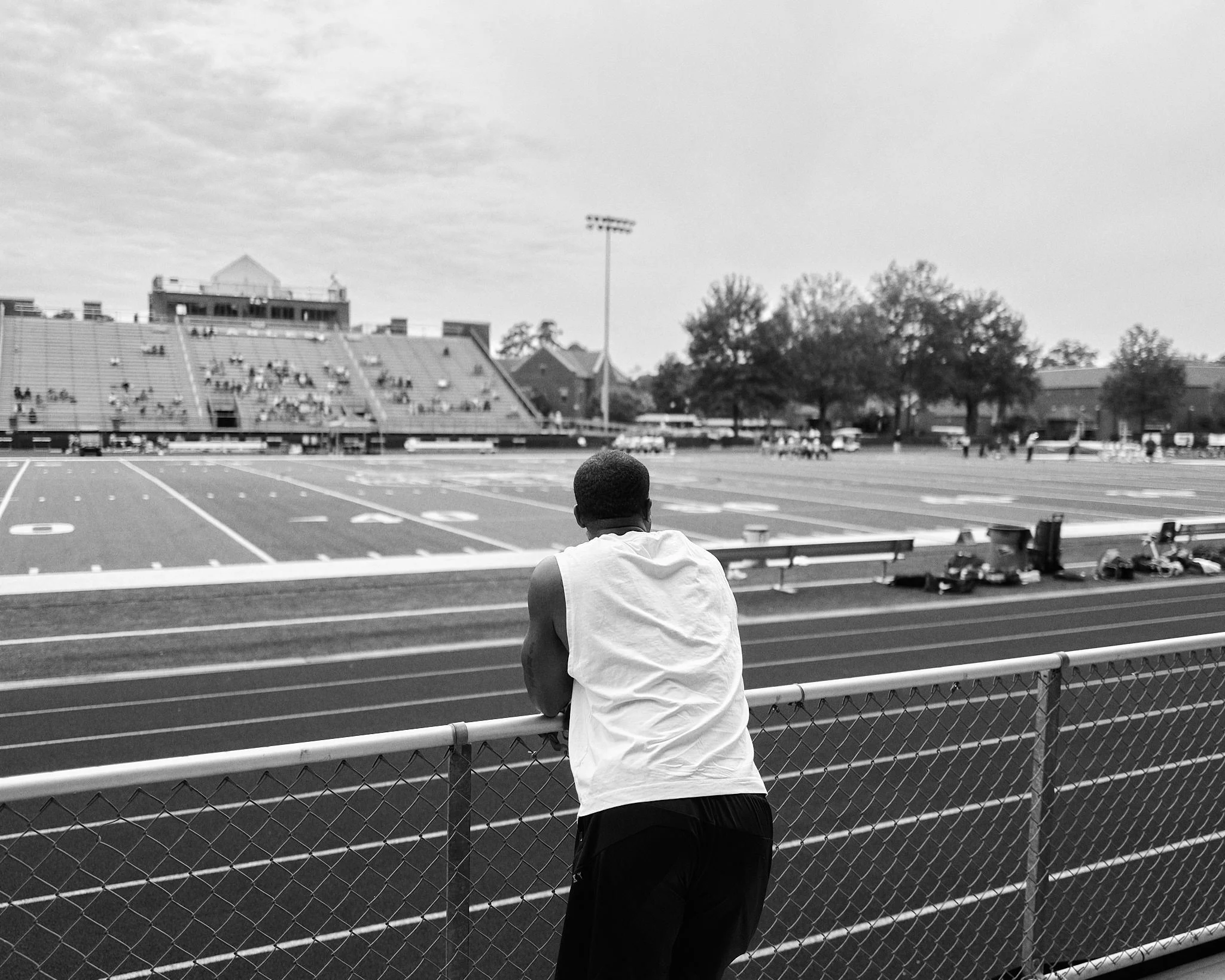 Devale Ellis leaning on a fence, watching a football field on a cloudy day.