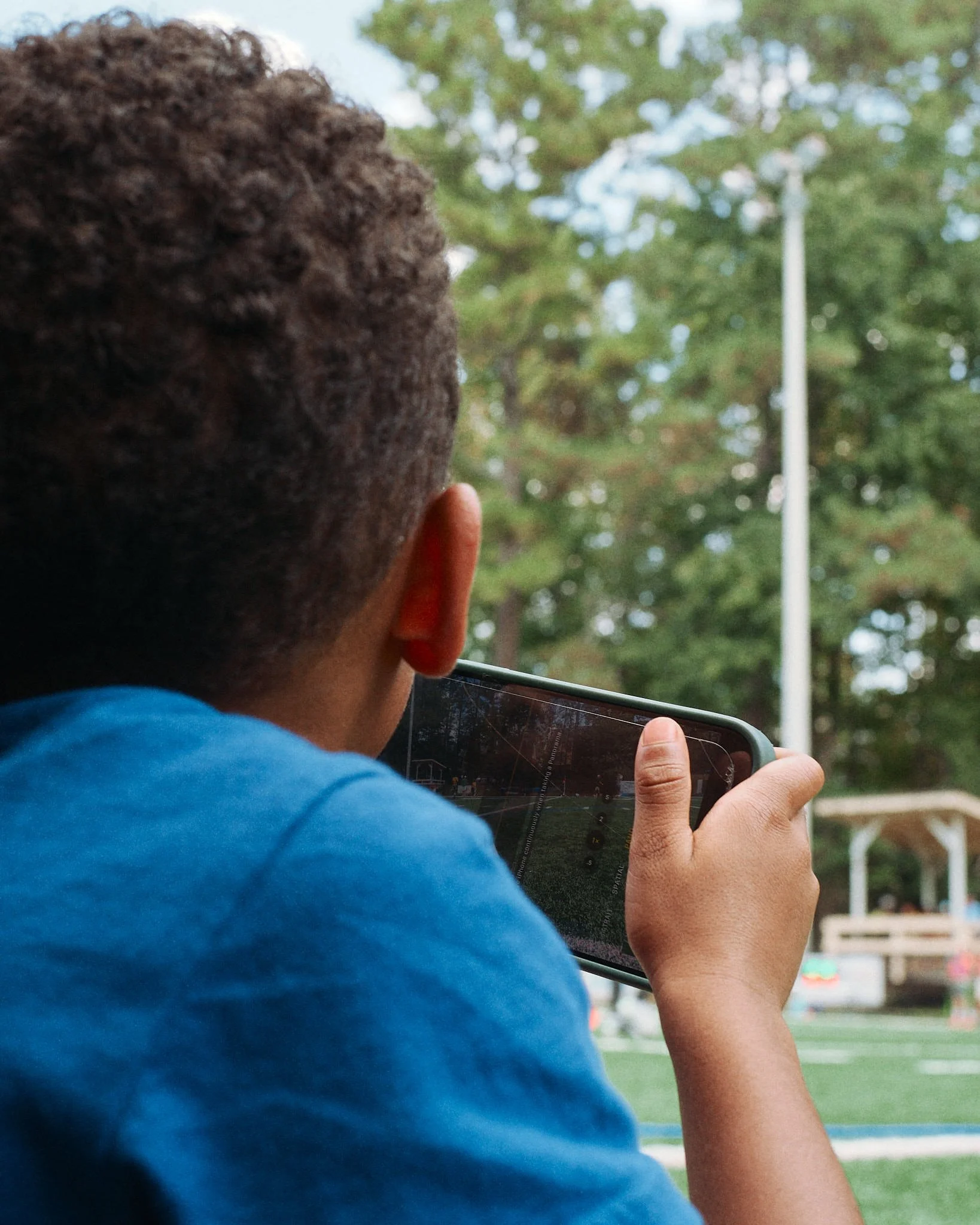 A boy with curly hair wearing a blue shirt is taking a photo with his smartphone outdoors near green trees and a playground.