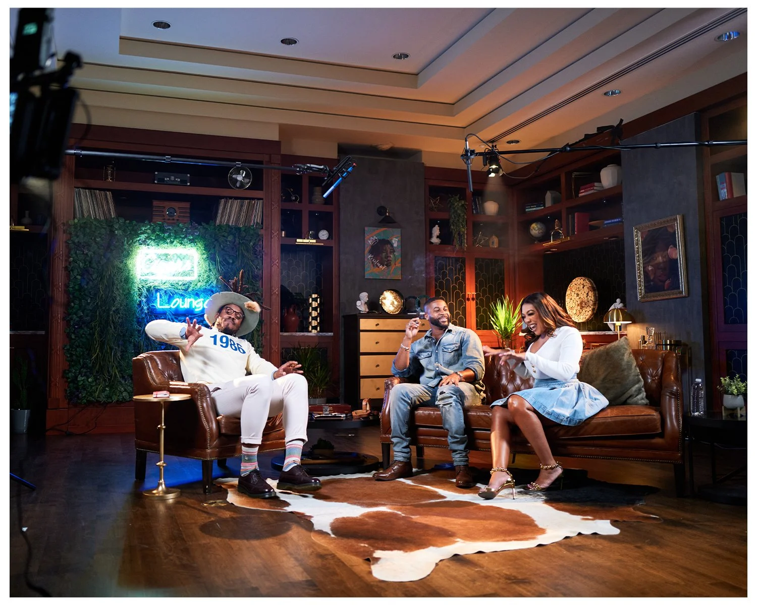 Three people sitting on leather couches in a cozy, well-decorated room, participating in an interview or talk show with professional film equipment overhead, a neon sign reading 'Lounge,' and various decorative items on shelves.