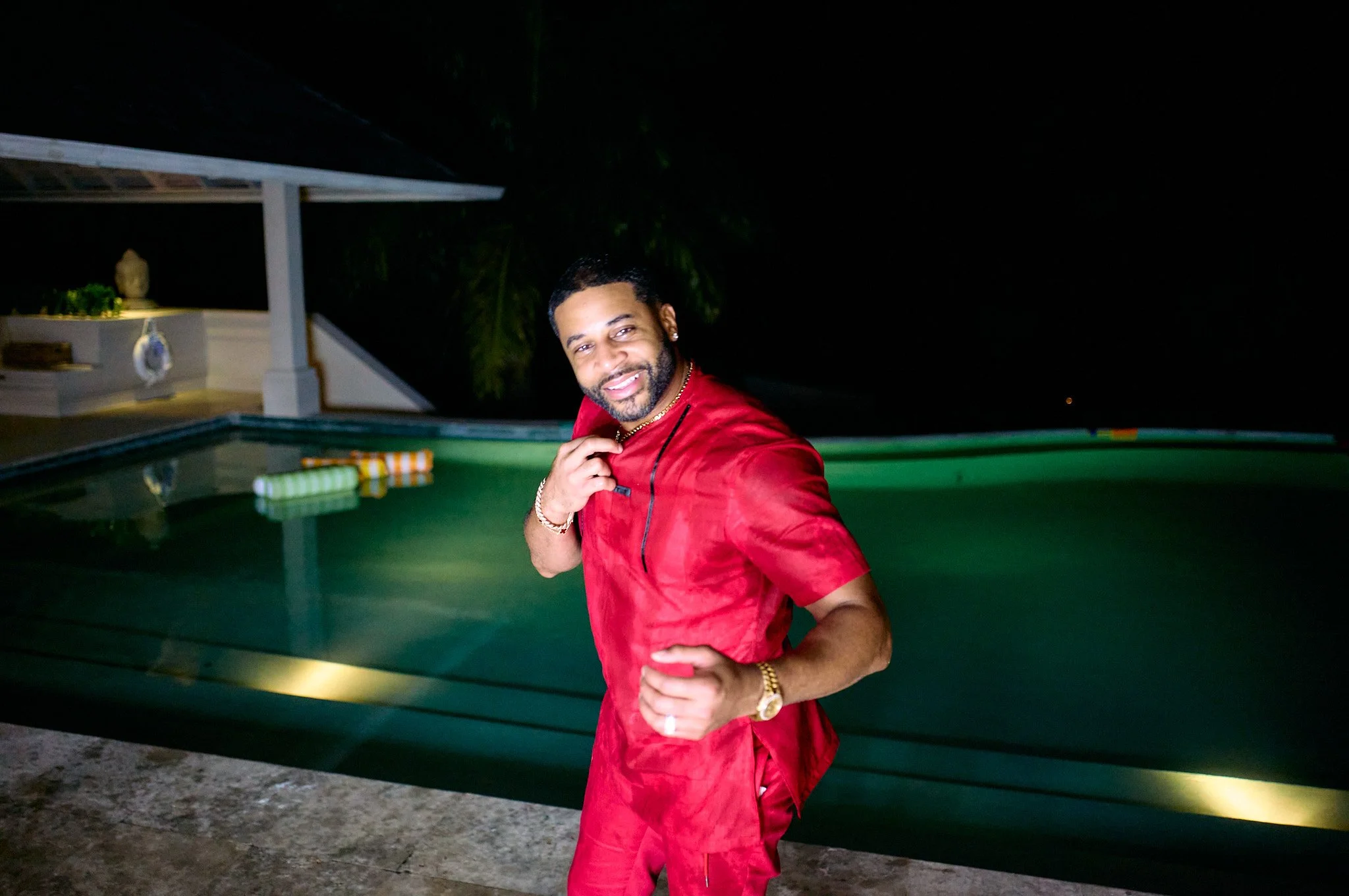 A man in a red outfit standing by an illuminated swimming pool at night, smiling and posing for the camera.