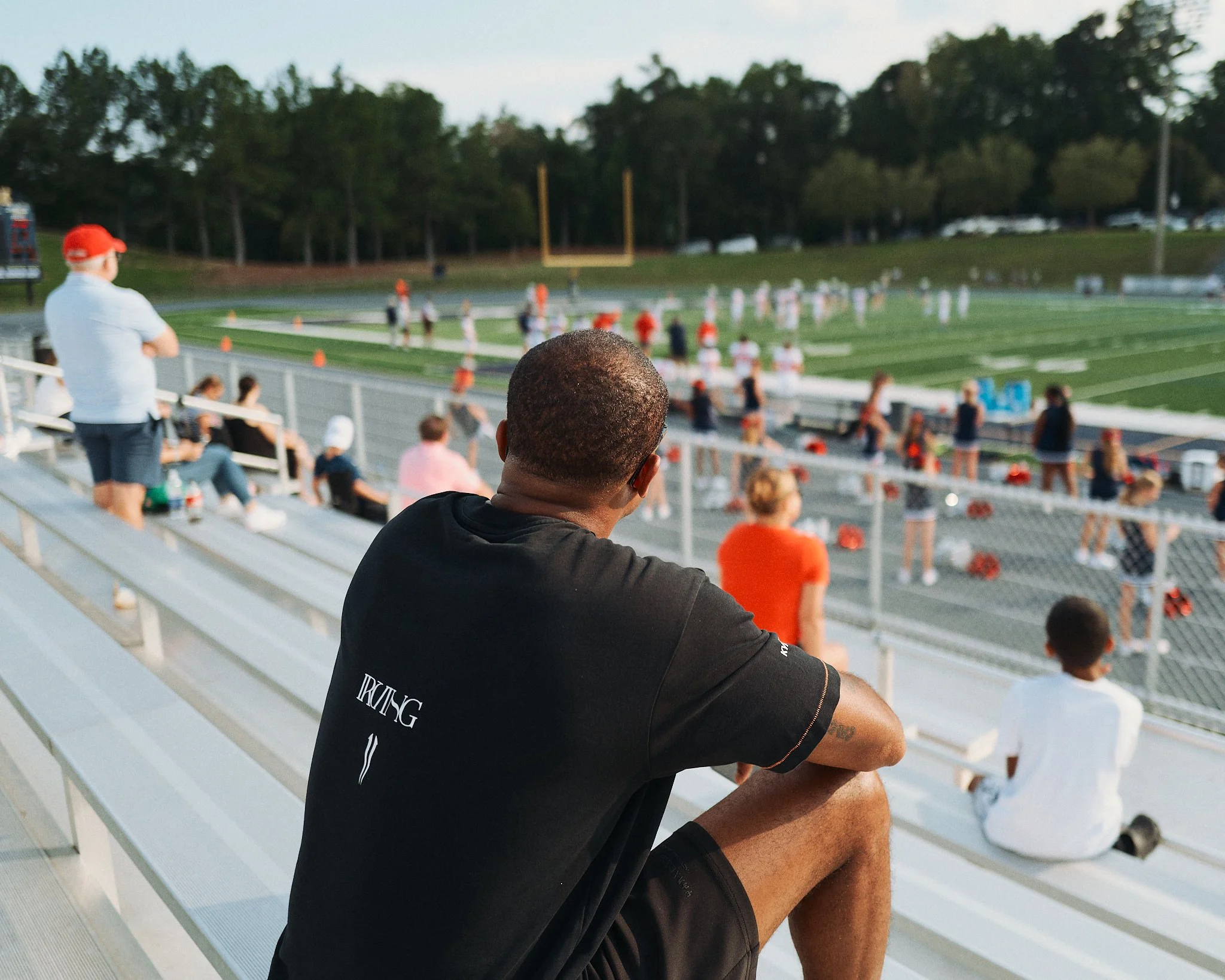 Devale Ellis sitting on bleachers watching a football game on the field with children and adults in attendance.