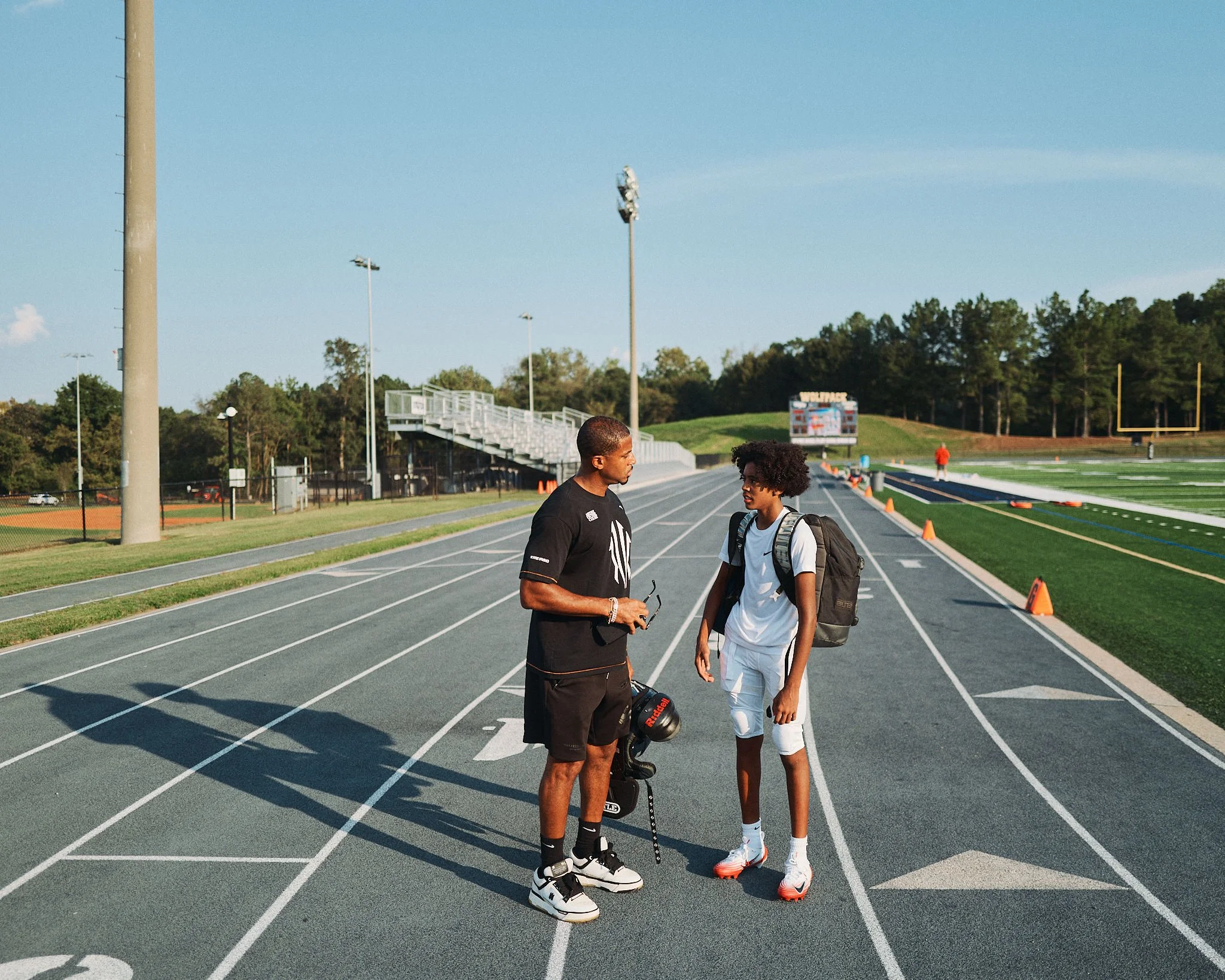 Two young men standing and talking on an outdoor track field in the late afternoon. One is holding sunglasses and a helmet, wearing athletic gear, and the other has a backpack and is dressed in white sportswear. They are engaged in conversation under