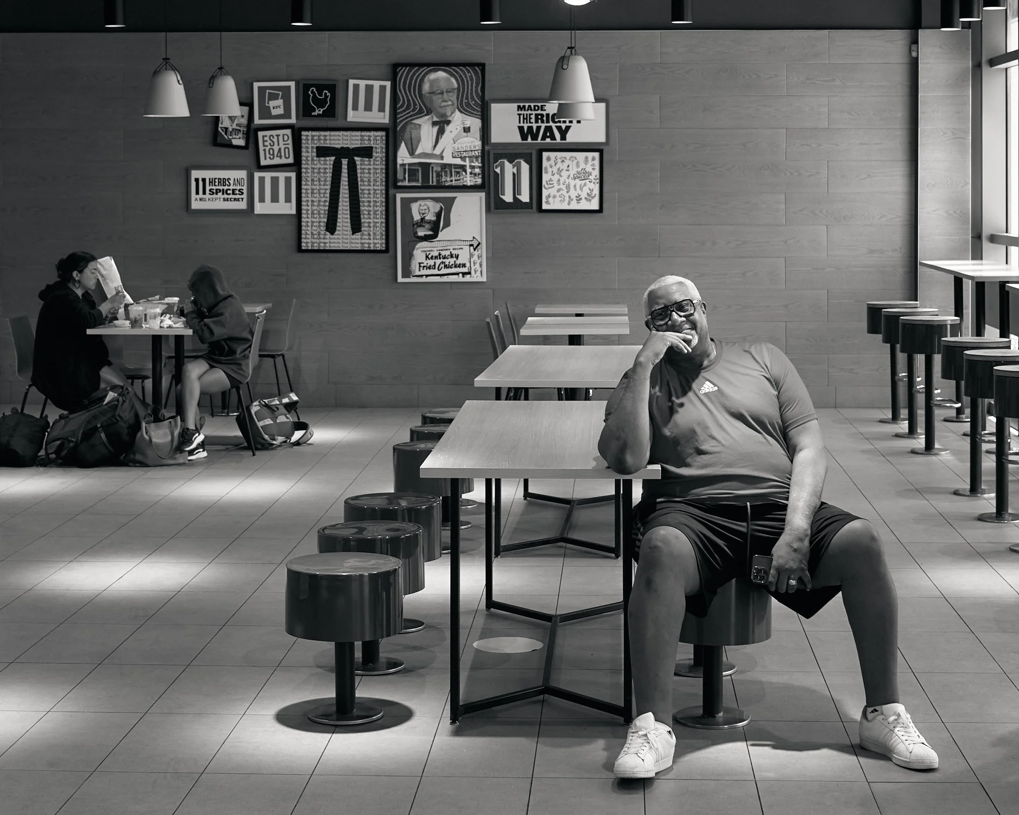 A man sitting at a table in a restaurant, smiling and resting his head on his hand, with two women seated at a table in the background eating. The restaurant has framed pictures and signs on the wall, including one reading "Made the Right Way."