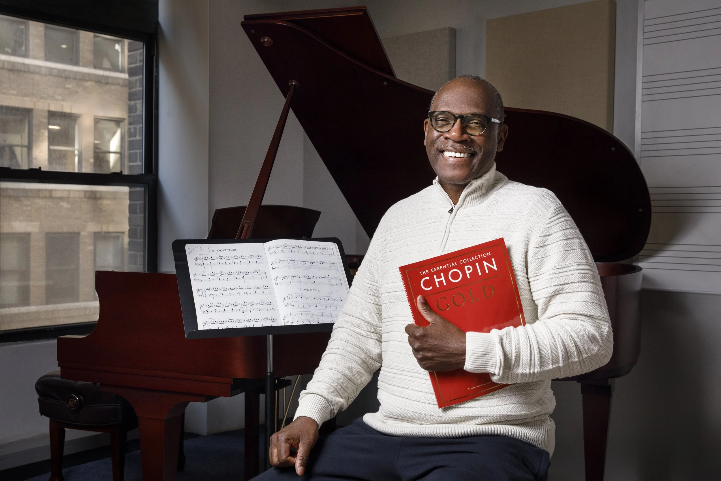 a portrait of a man smiling holding a red music book titled 'Chopin Gold' in a music studio with a grand piano and sheet music in the background.