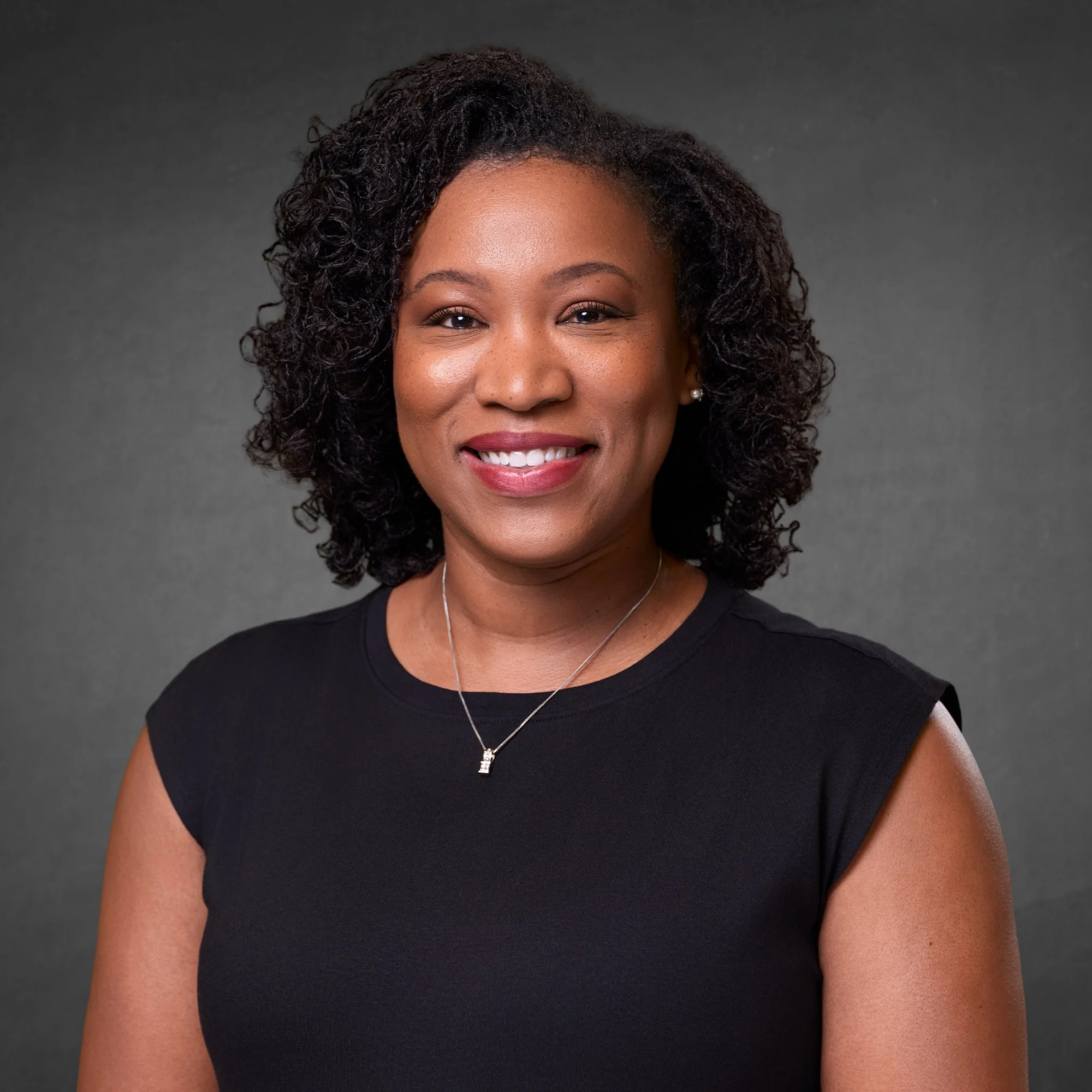 Professional headshot of a woman with, curly hair wearing a black top and a silver necklace, smiling against a gray background.