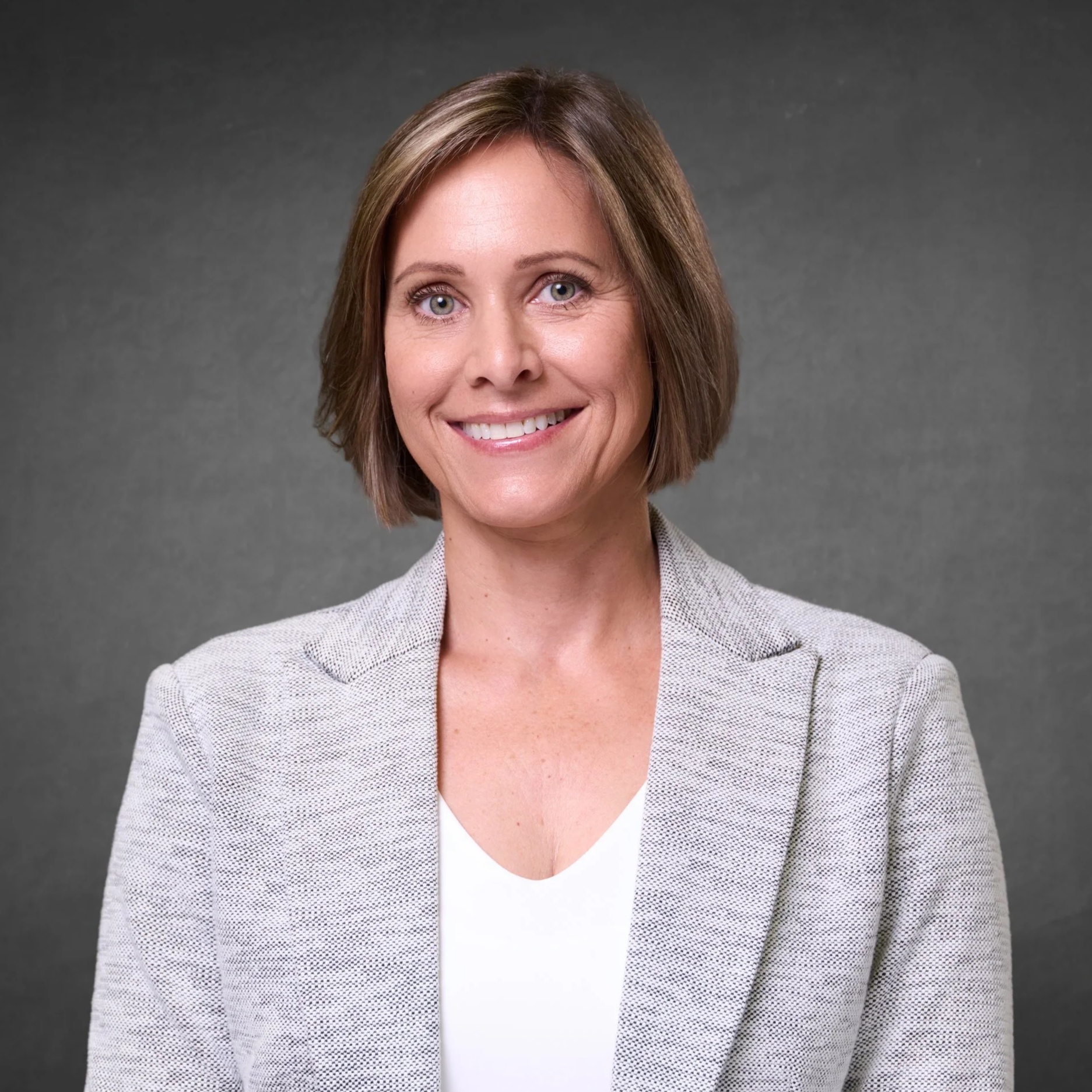 A smiling woman with short brown hair wearing a light gray blazer over a white top, posed against a plain gray background.
