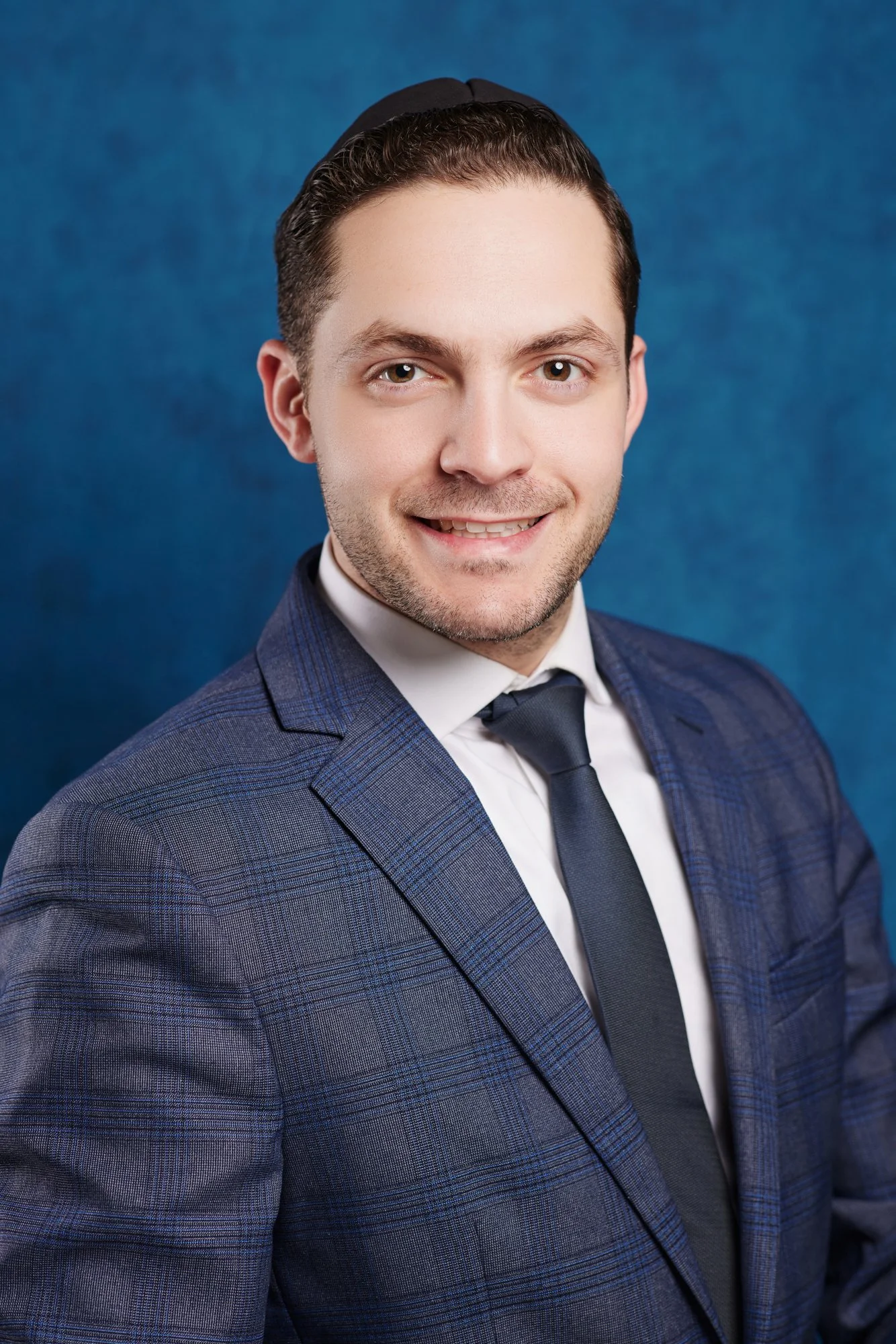 Headshot of a jewish young man with light skin, brown hair, wearing a dark suit and a tie, smiling against a blue background.