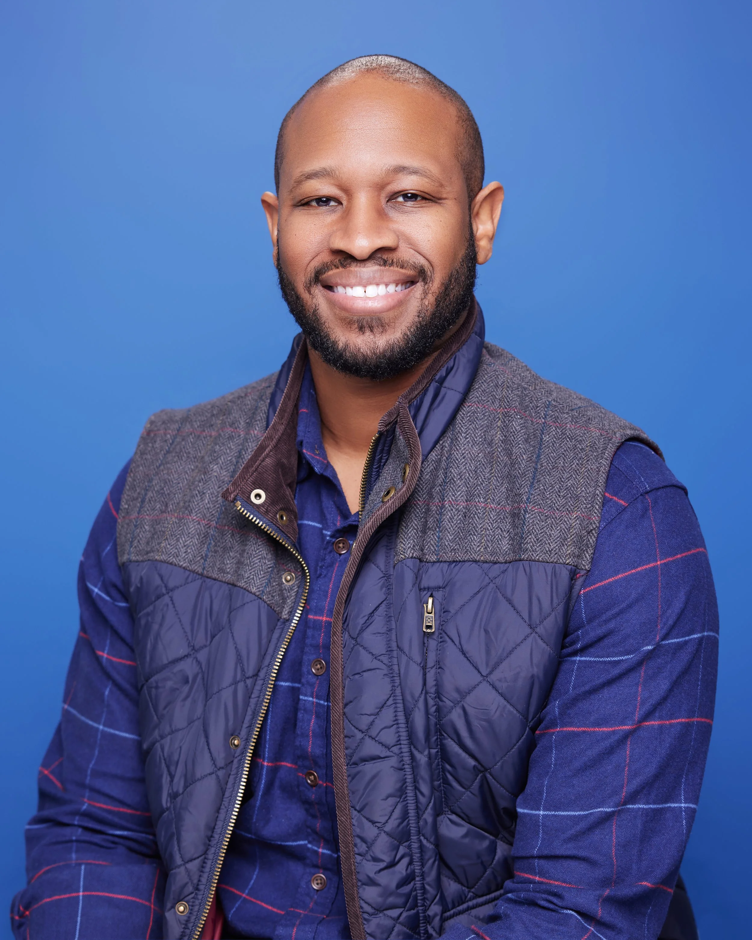A smiling man with a beard and short hair, wearing a blue button-up shirt and a gray and blue vest, posed against a plain blue background.