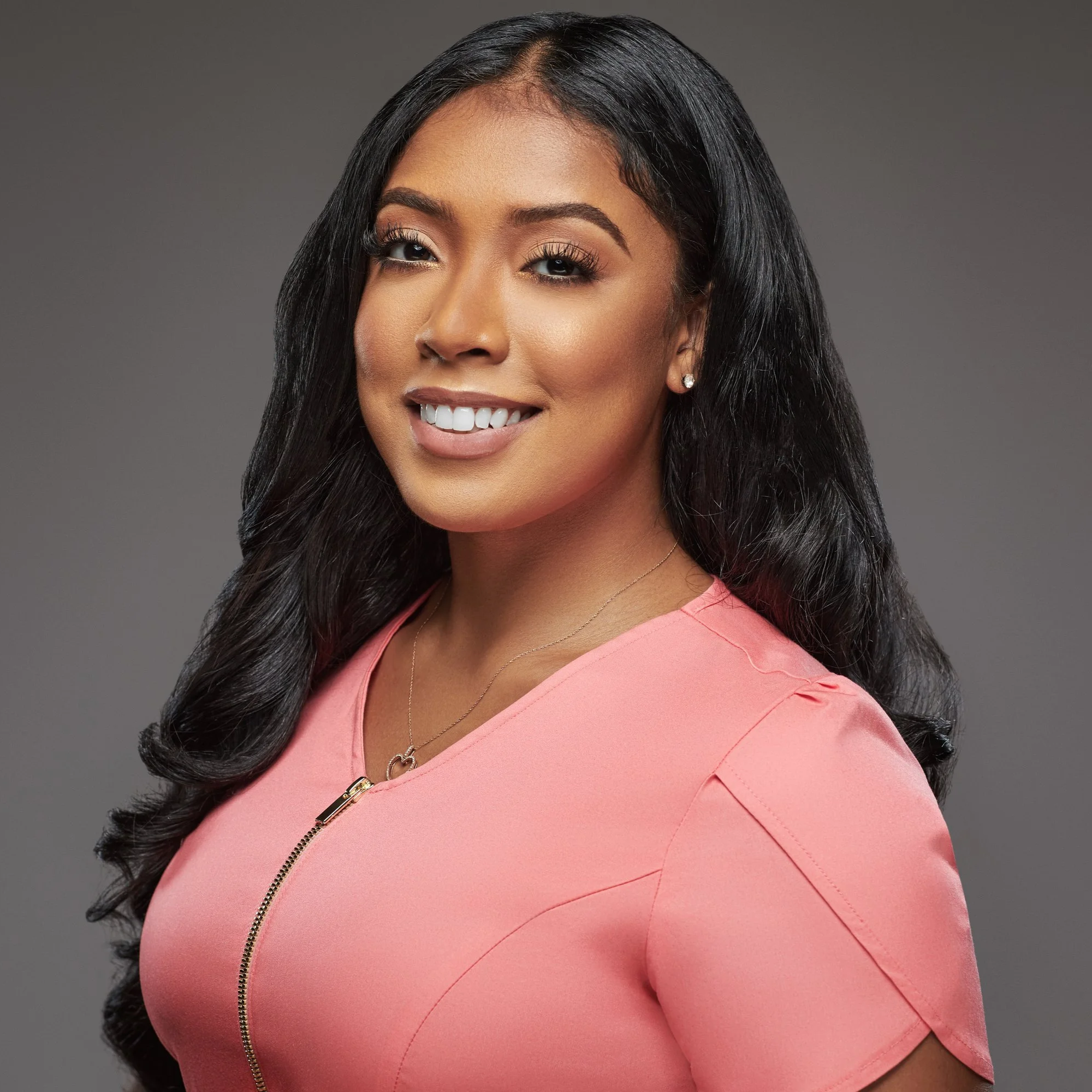Portrait of a smiling woman with long black hair wearing a pink top and jewelry against a gray background.