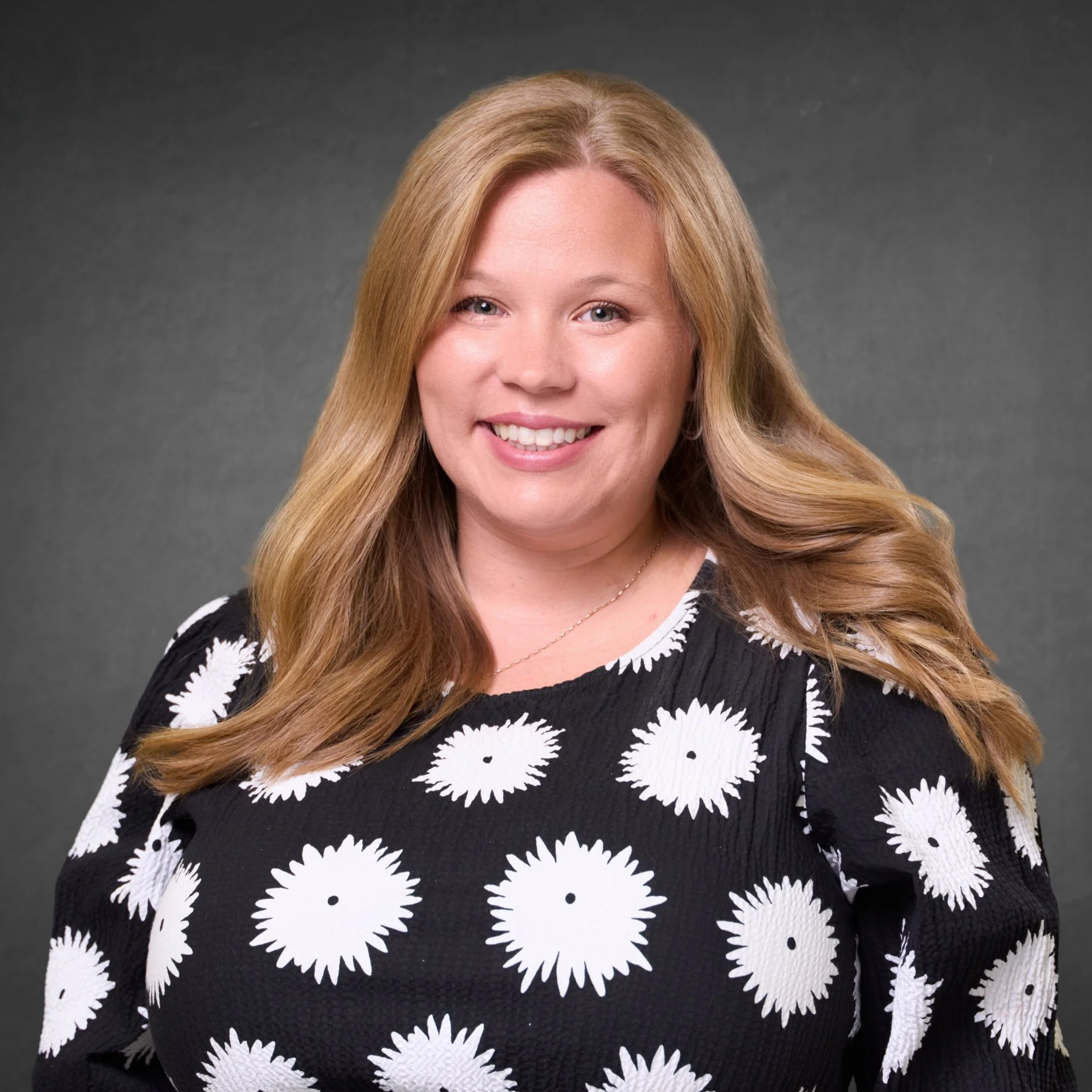 A woman with long wavy blonde hair, smiling, wearing a black dress with white daisy patterns, and standing against a gray background.