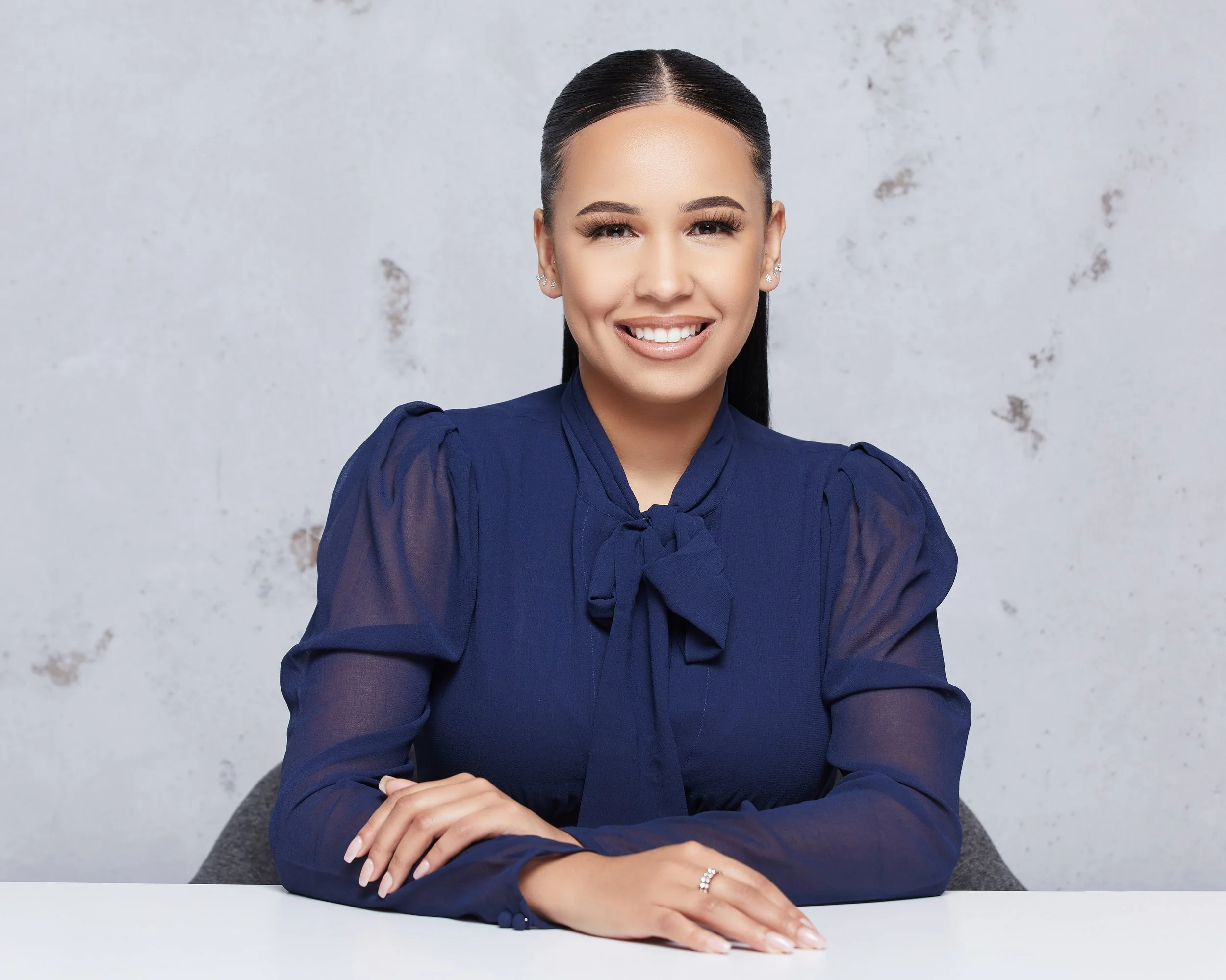 A woman with long dark hair pulled back, wearing a navy blue blouse with a bow tie, smiling and sitting at a table with arms crossed.