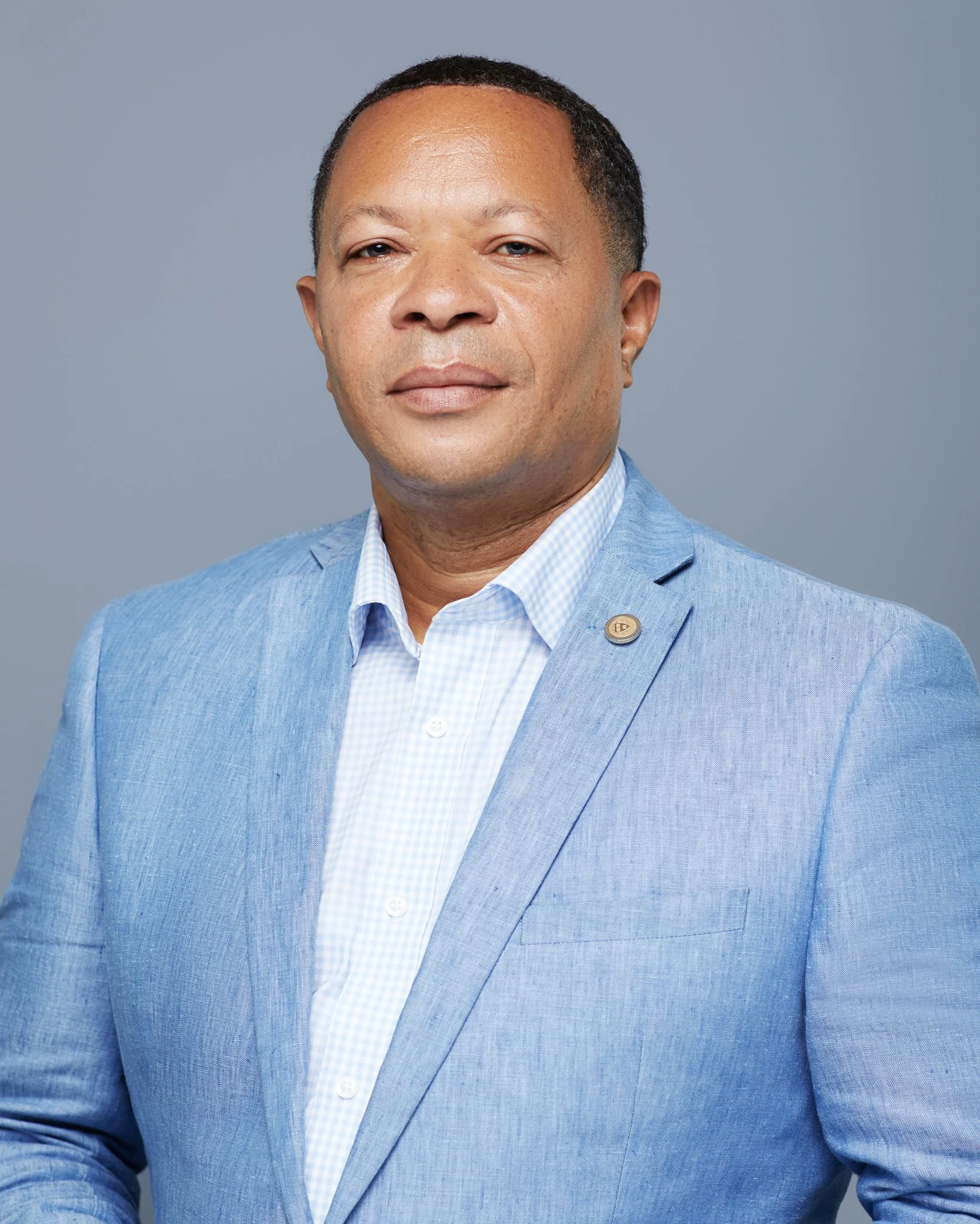 A man in a light blue suit and white checkered shirt is posing against a gray background.