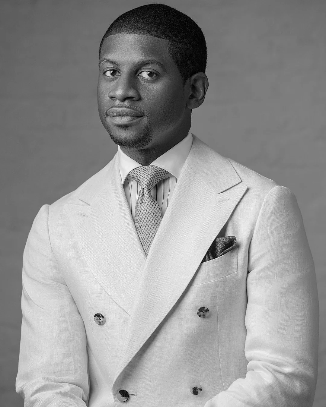 Black and white portrait of a man in a light-colored suit with a patterned tie and pocket square, looking at the camera with a neutral expression.