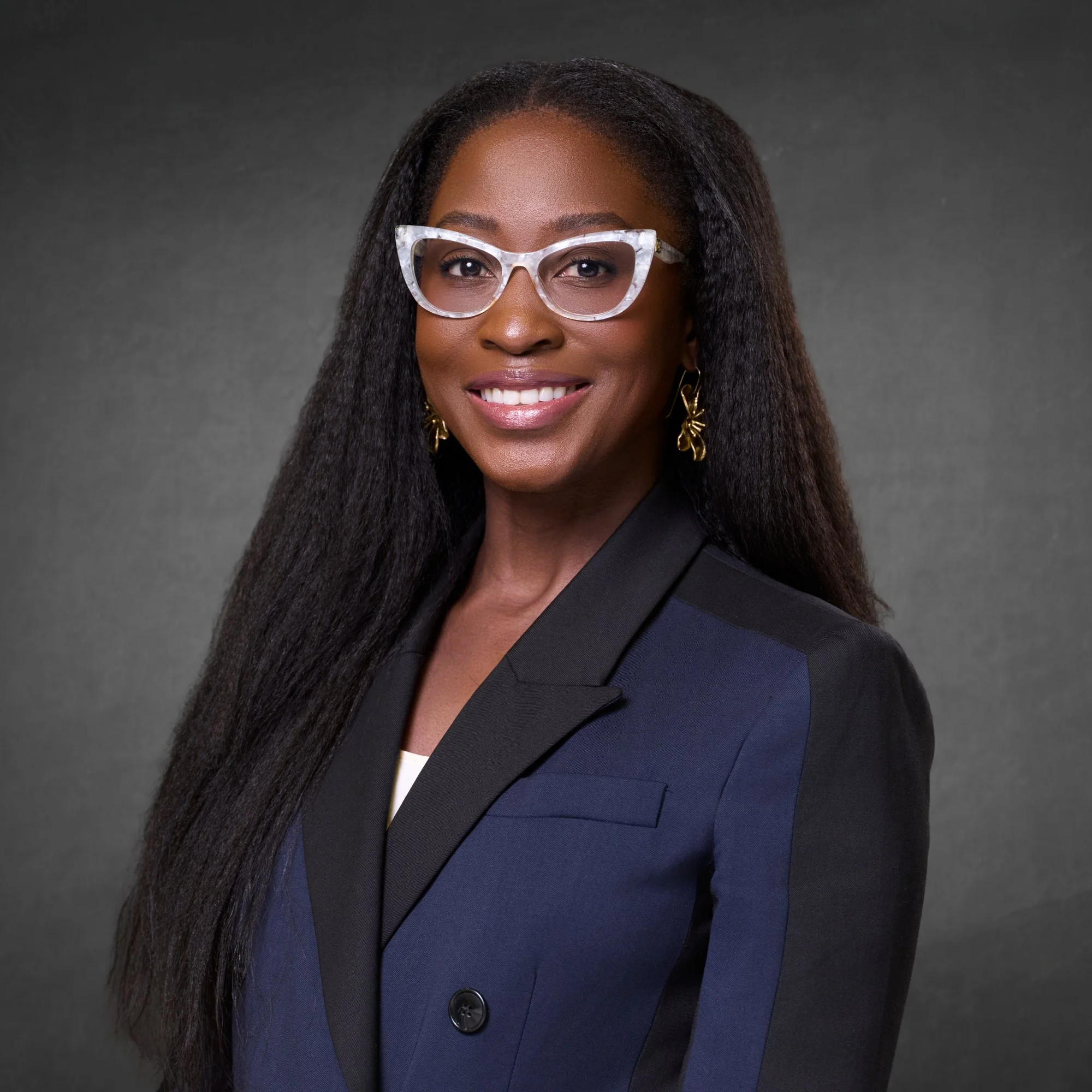 A woman with long black hair wearing glasses, gold earrings, and a dark blazer, smiling against a gray background.