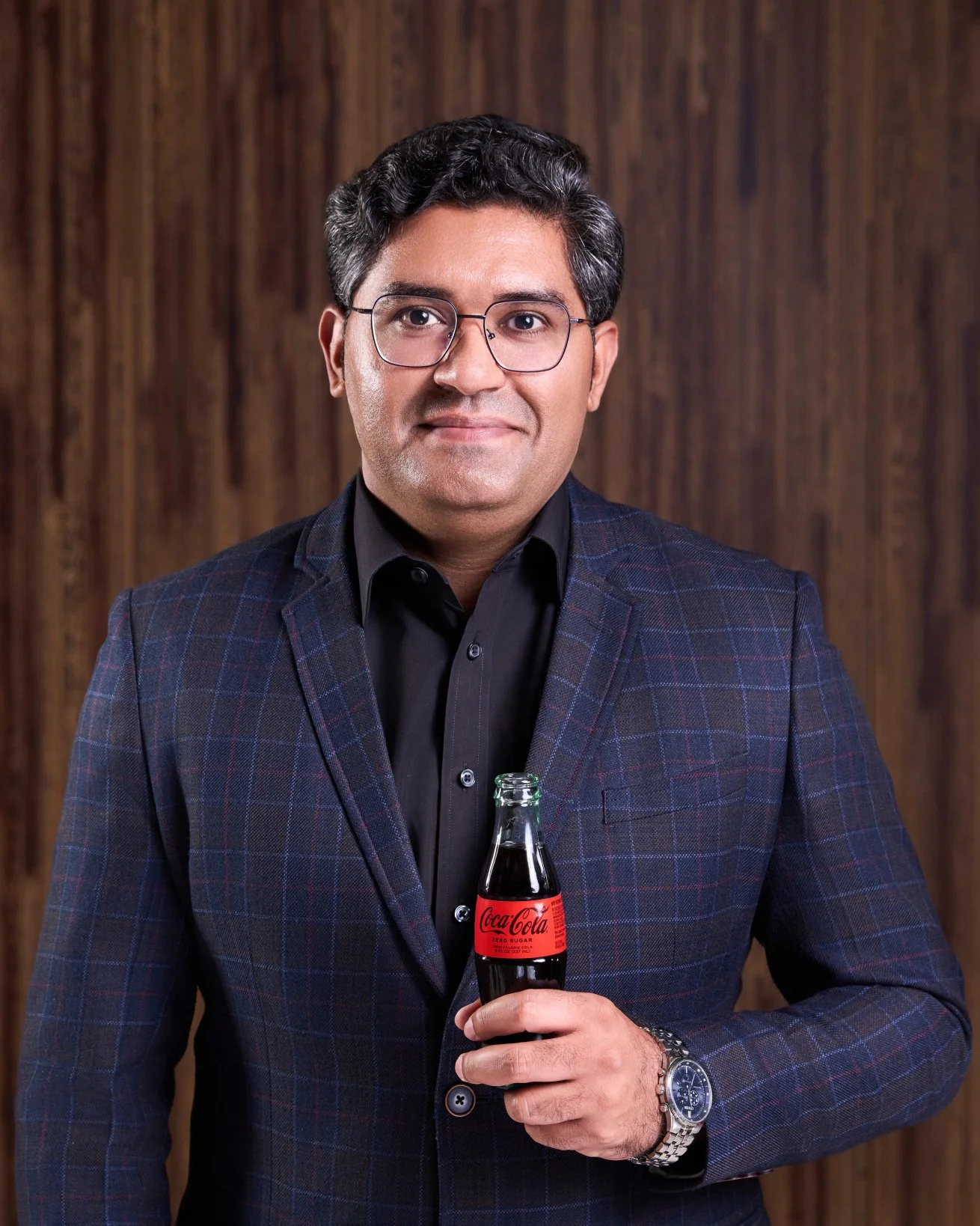 Portrait of Man in glasses and dark blazer holding a glass bottle of Coca-Cola, standing in front of a wooden background by Matt Ellis