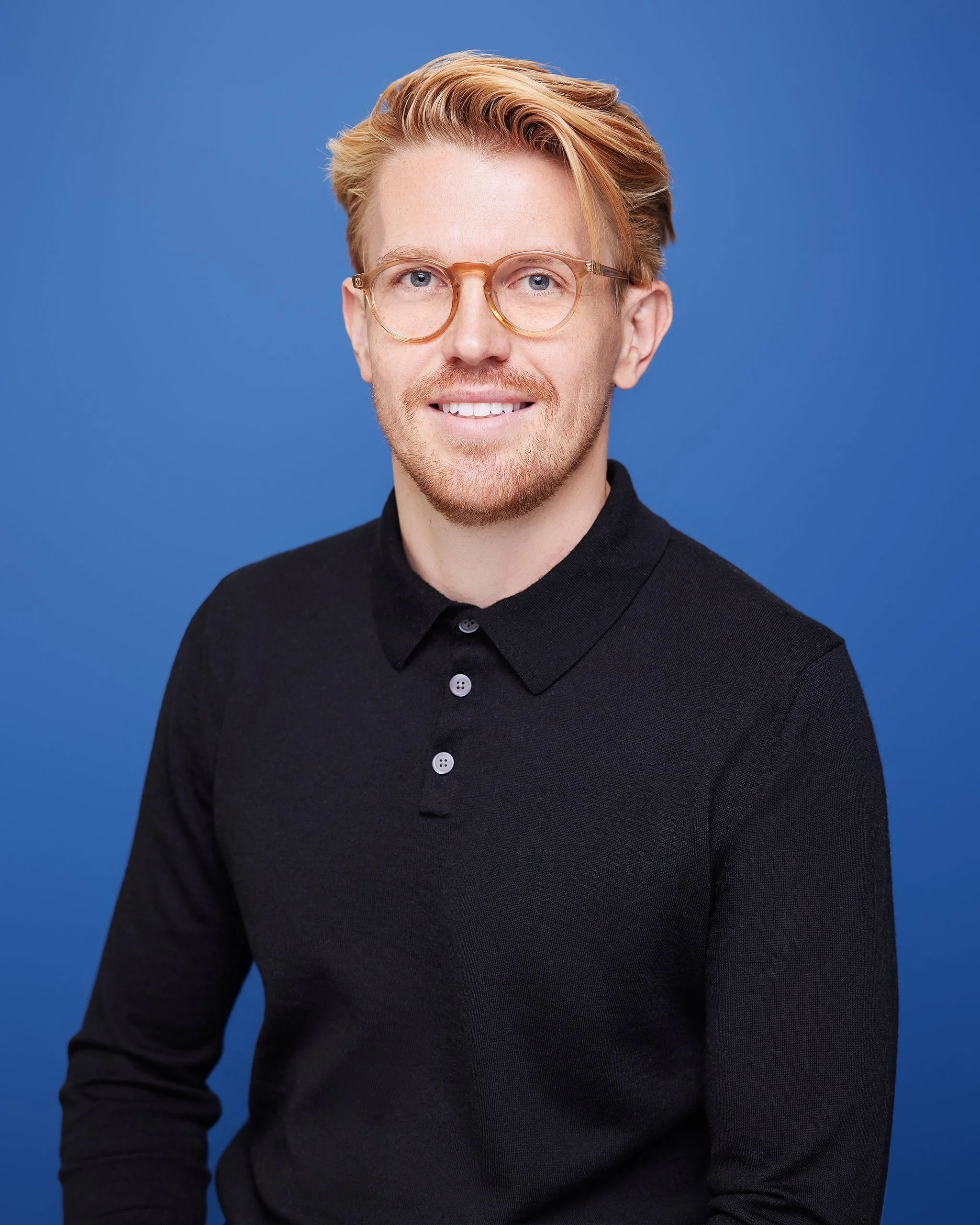 A young man with blond hair, beard, glasses, wearing a black polo shirt, smiling against a blue background.