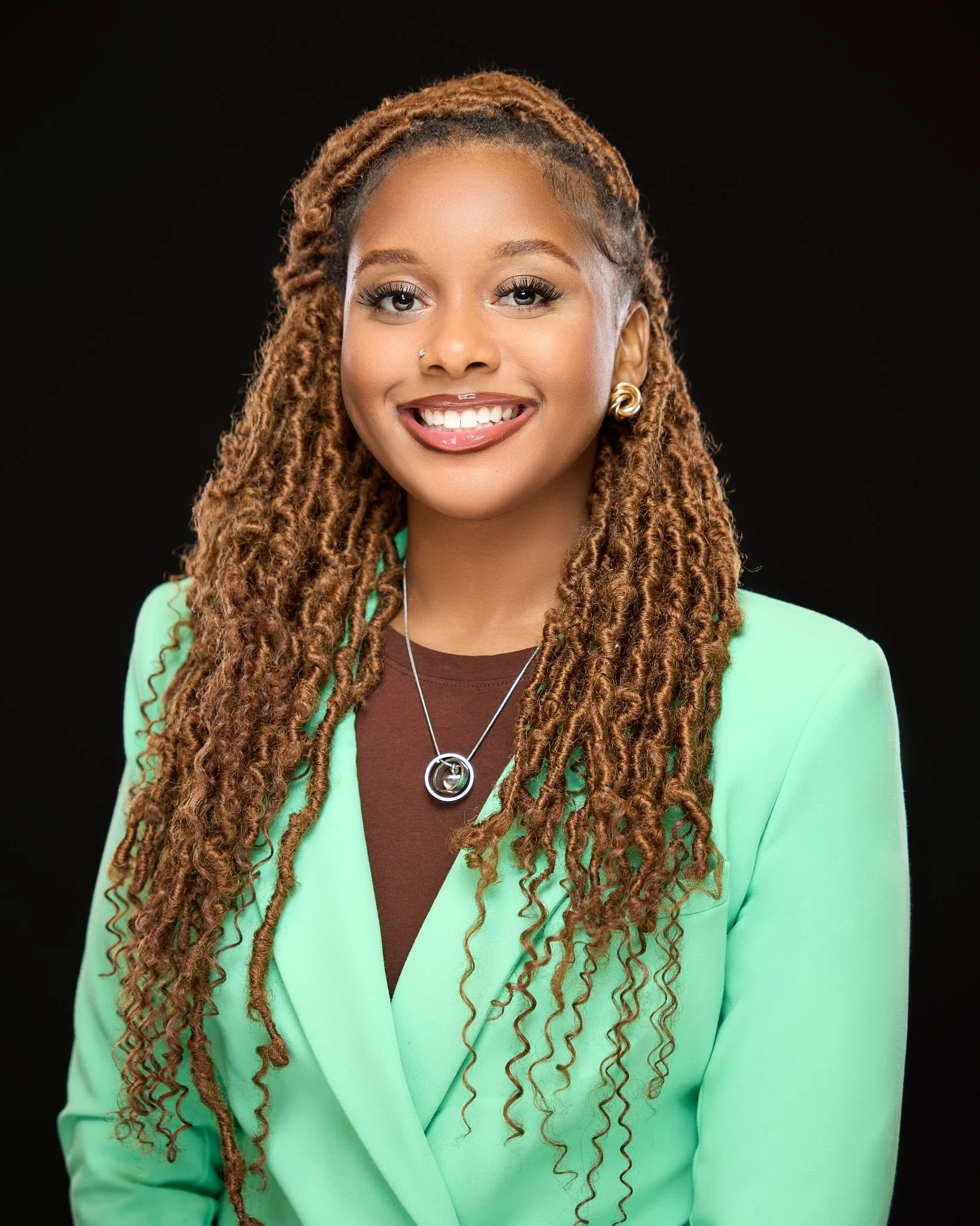 Portrait of a young woman with caramel-colored dreadlocks, smiling, wearing a mint green blazer, brown top, and silver jewelry, against a black background.