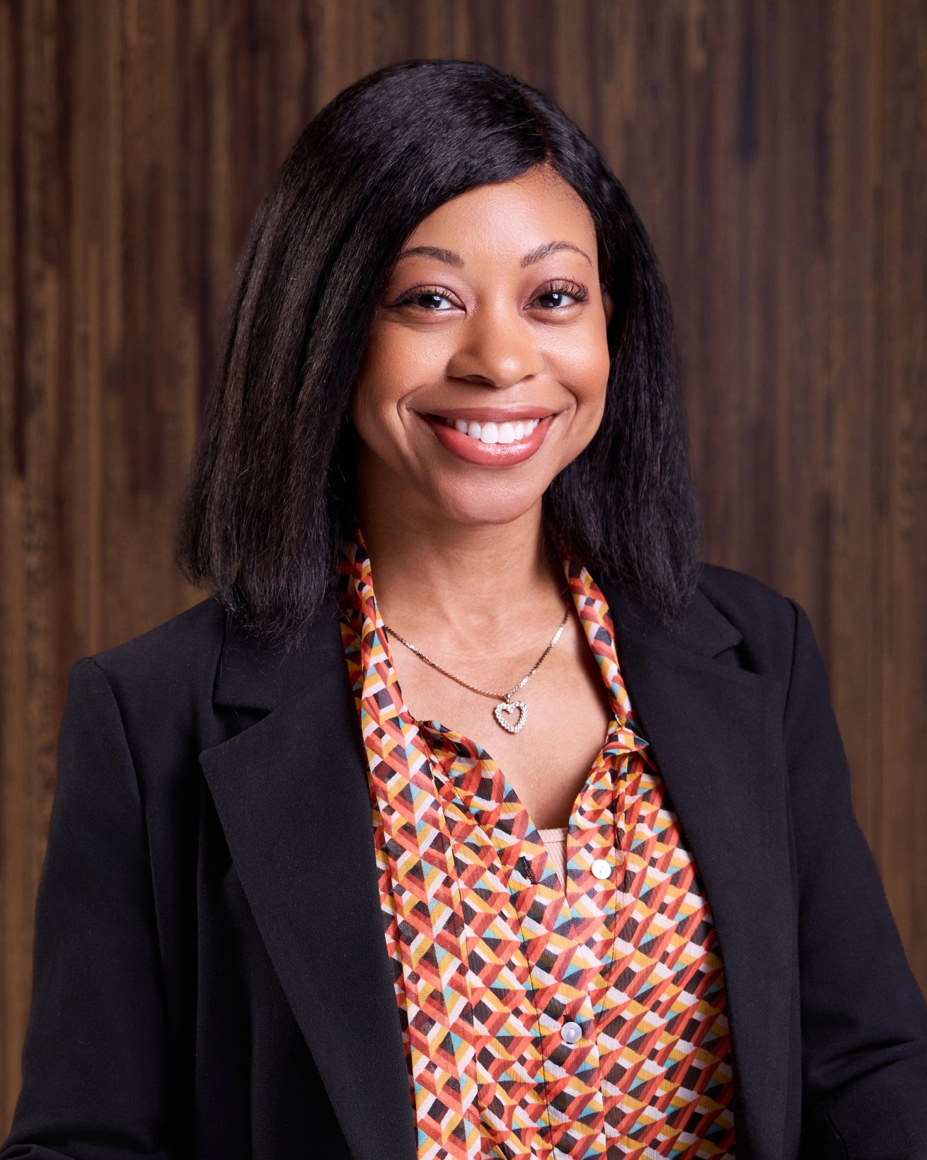 A woman with medium-length black hair, smiling, wearing a black blazer, colorful patterned blouse, and a heart-shaped necklace, standing against a wooden background.
