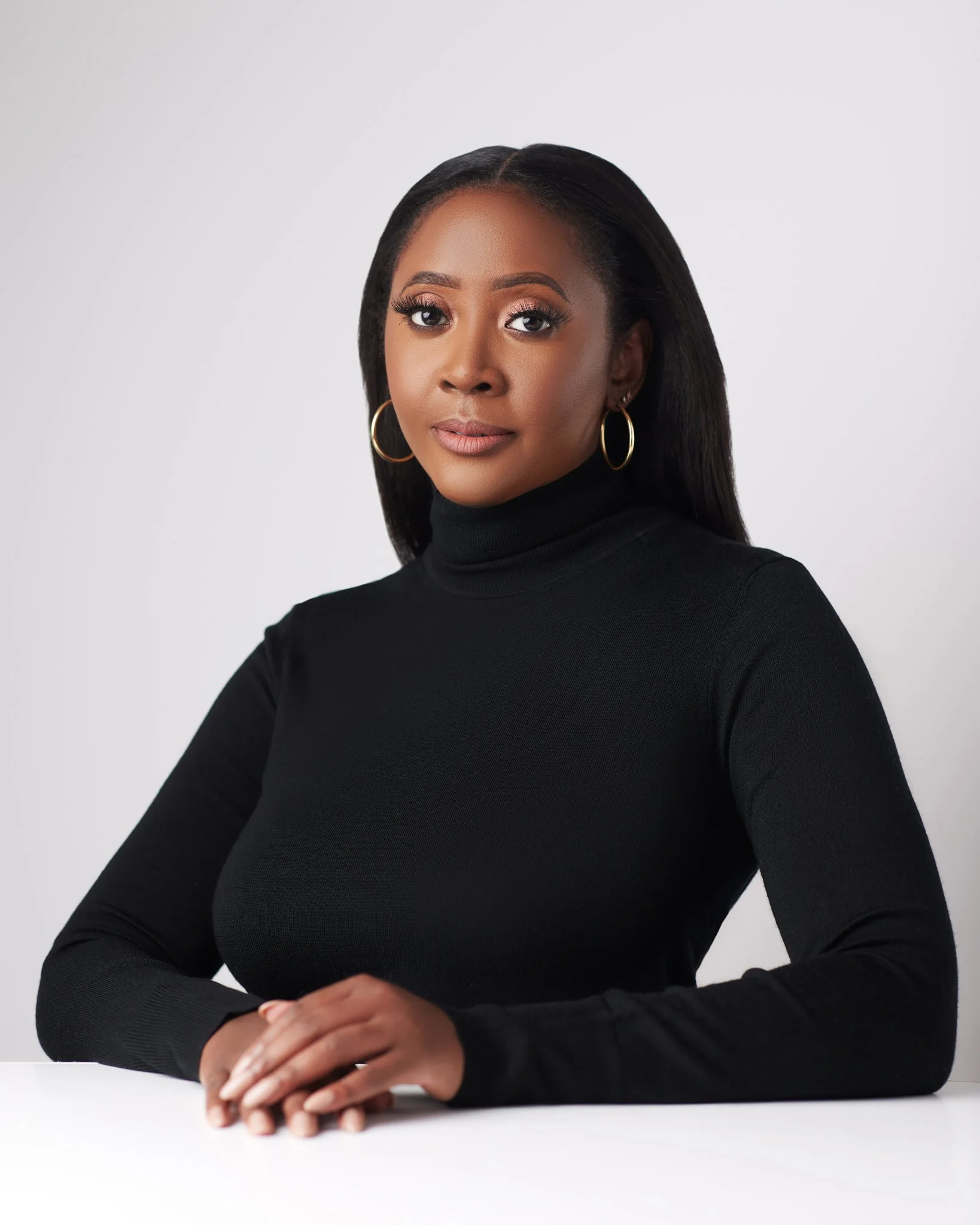Portrait of a woman with long dark hair, gold hoop earrings, wearing a black turtleneck, sitting with hands clasped on a white surface, against a plain gray background.