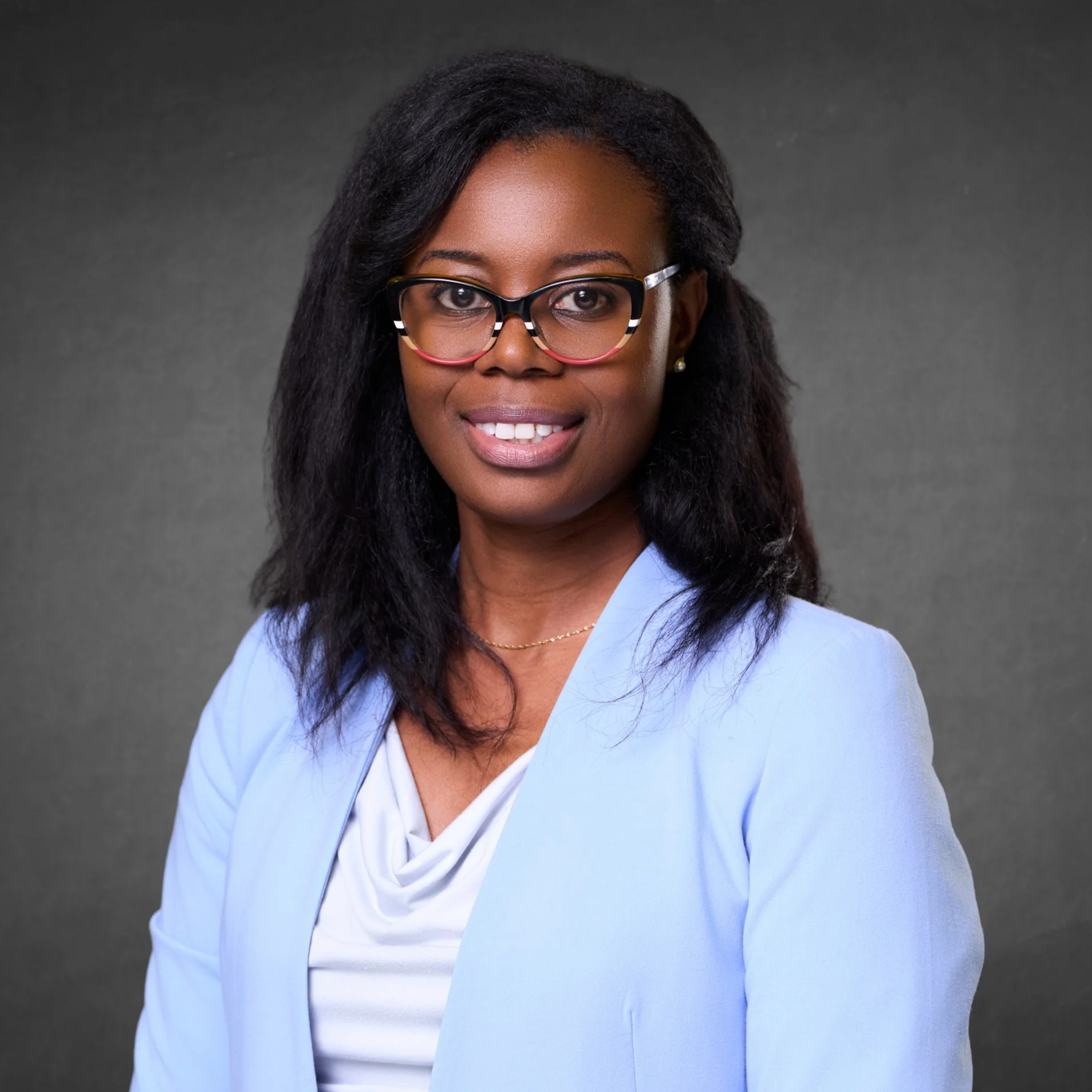 Professional woman with glasses smiling, wearing a light blue blazer and white blouse against a dark gray background.