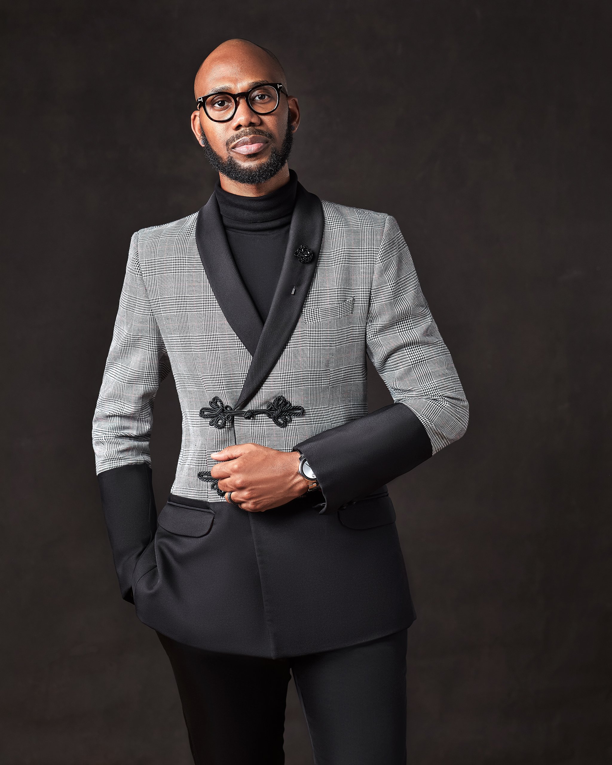 A man in a checked blazer with black satin lapel, black cuffs, and black buttons, wearing glasses, a black turtleneck, and black pants, standing against a dark background.