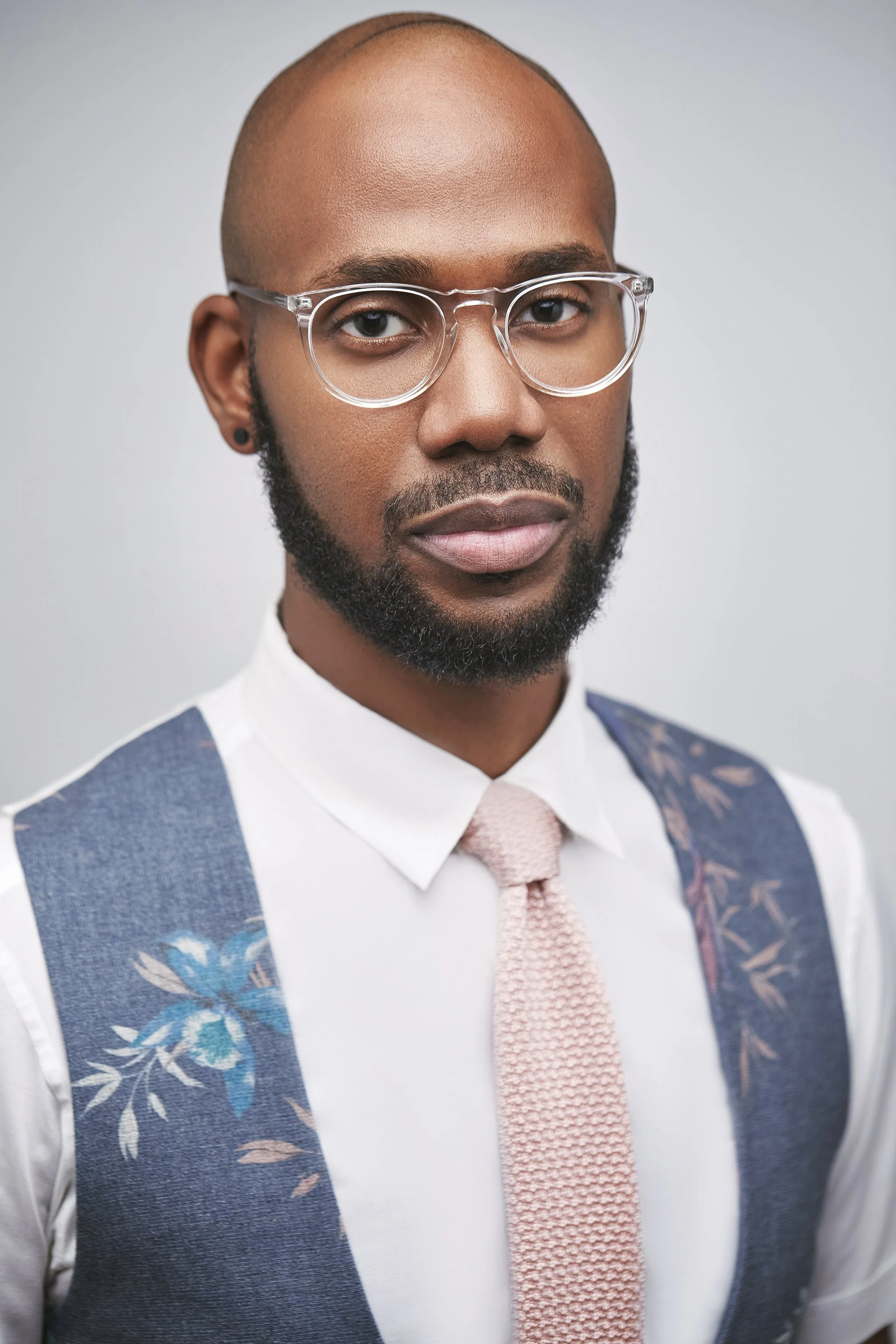 A man with a shaved head, glasses, and beard wearing a white shirt, pink tie, and a floral vest, against a plain background.